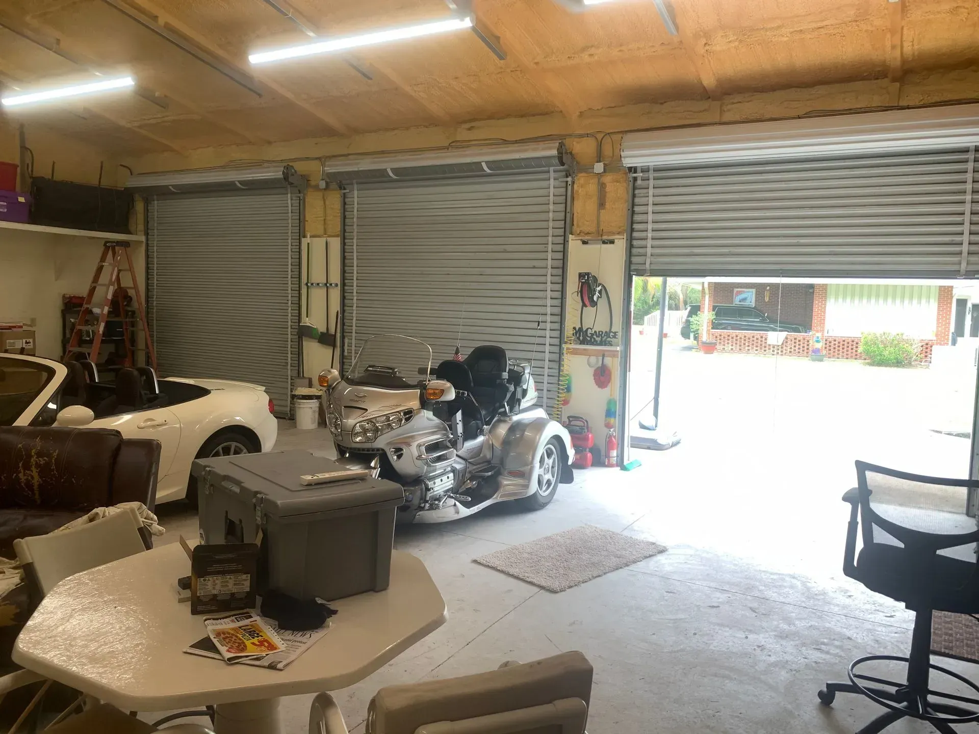 Garage interior with a trike motorcycle, car, table, and two closed garage doors. One door is open.