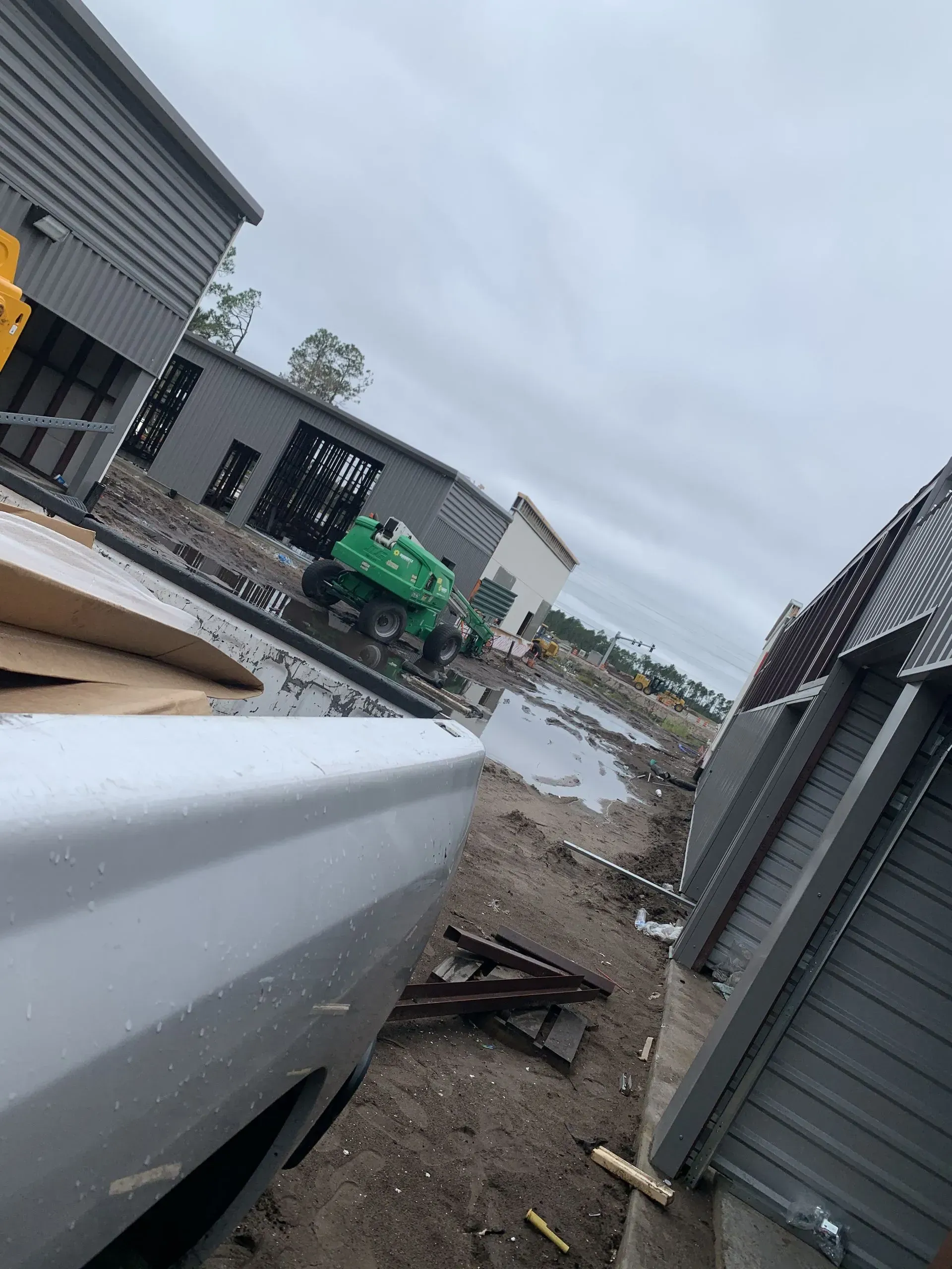 Construction site with buildings, truck, and a green lift. Overcast sky, muddy ground.