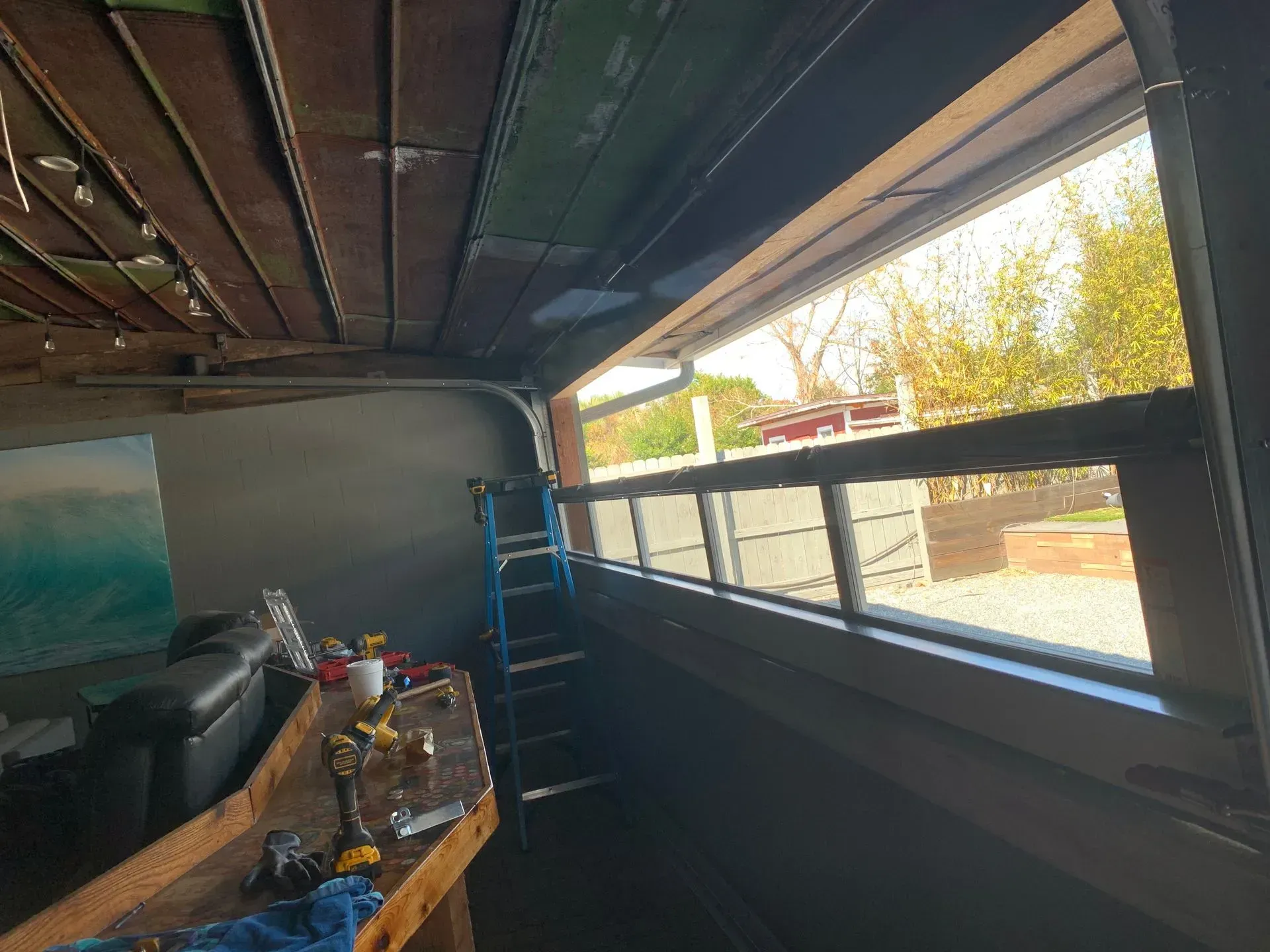 Interior view of a garage with a large window. A ladder, tools, and a partial view of the outside are visible.