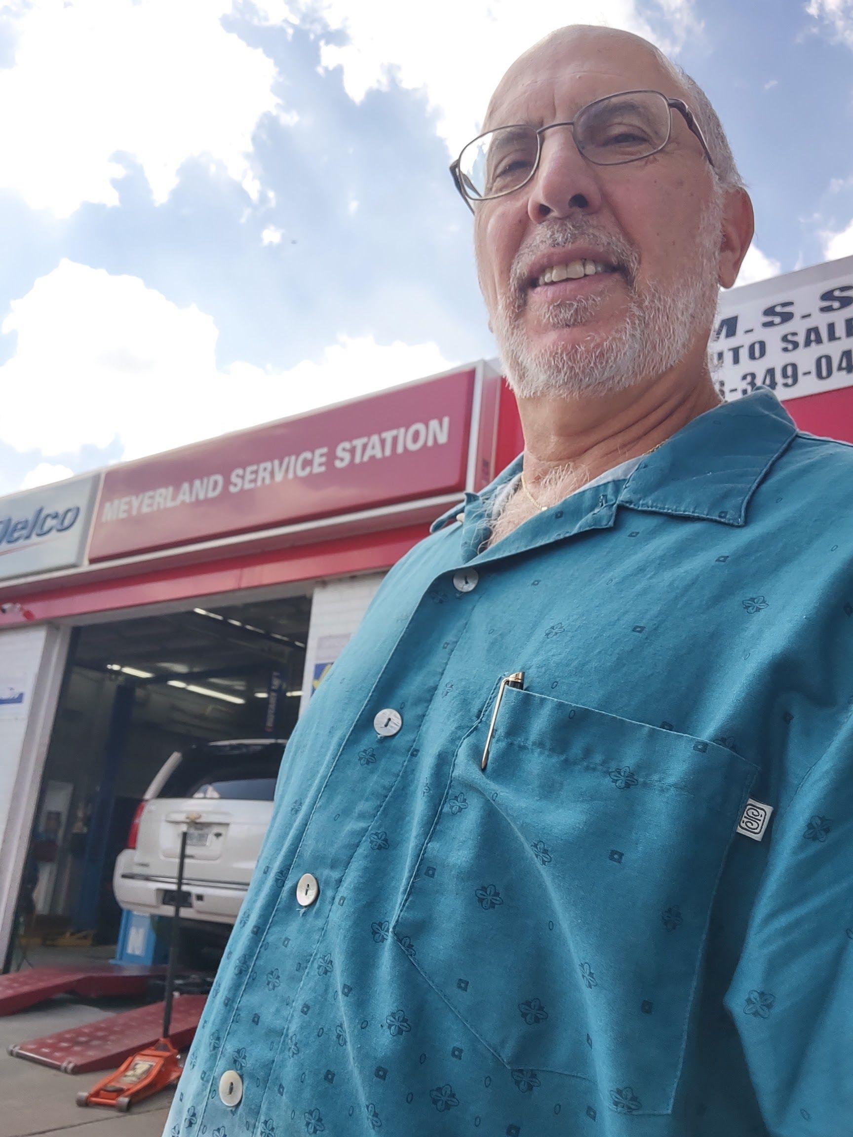Man in teal shirt in front of a service station with a car inside. Cloudy sky overhead. | Meyerland Service Station