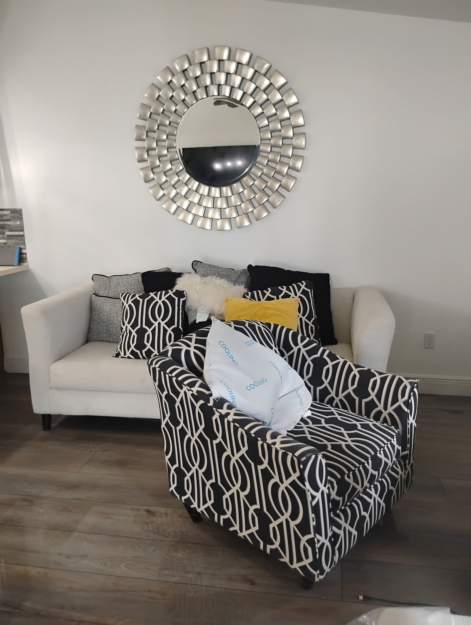 Living room with a white sofa and patterned chair, round silver mirror, and decorative pillows.