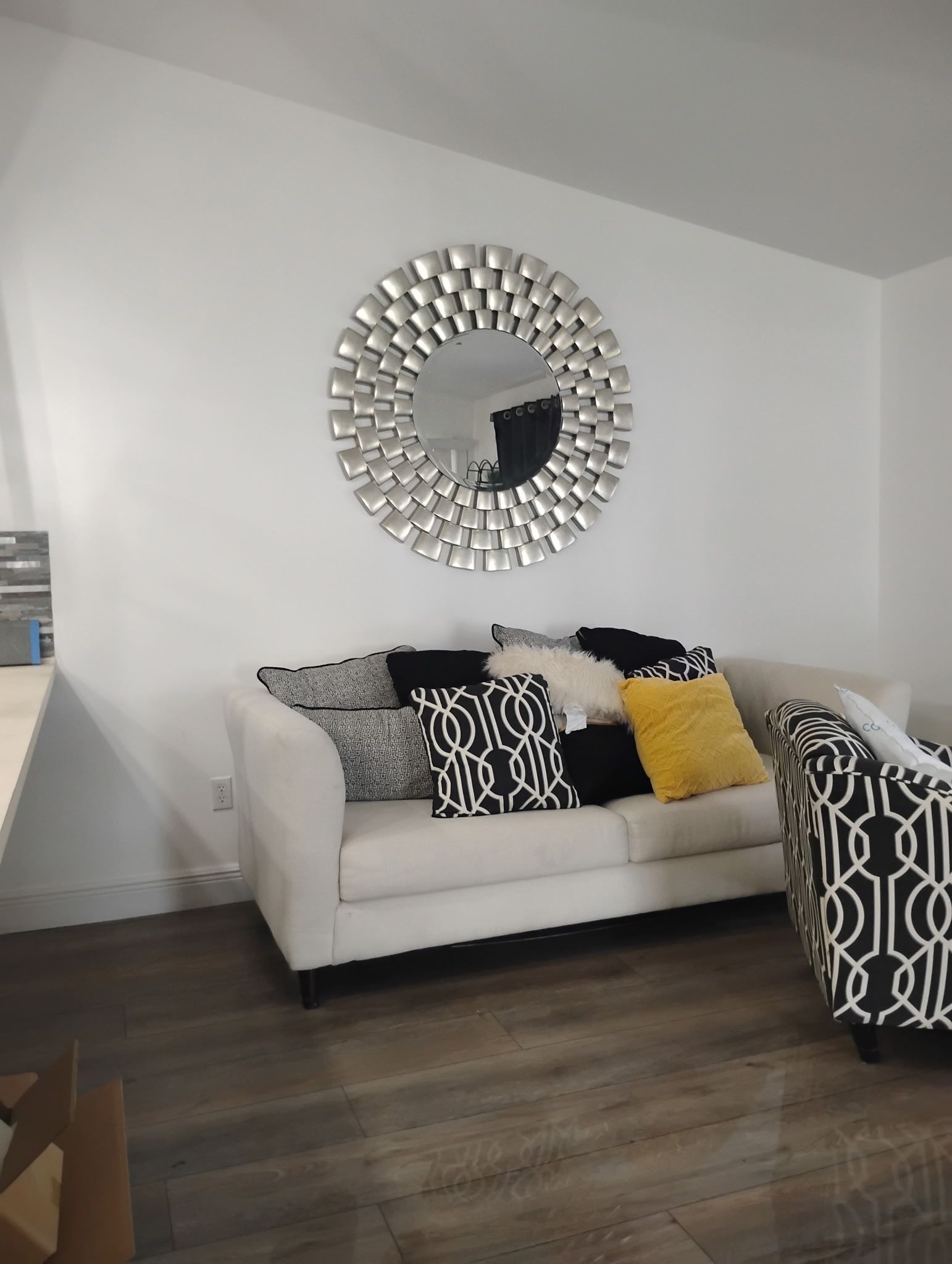 A white sofa with decorative pillows below a round silver mirror on a white wall.