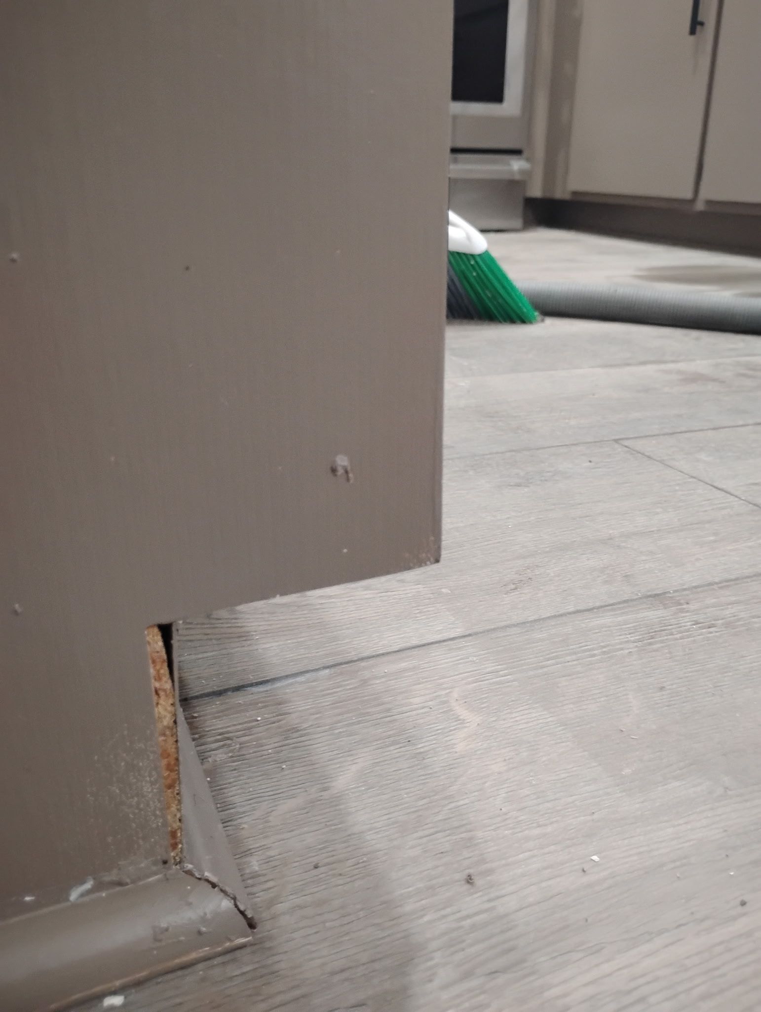 Close-up of a gray cabinet with a visible baseboard. The floor is light gray with a green broom in the background.