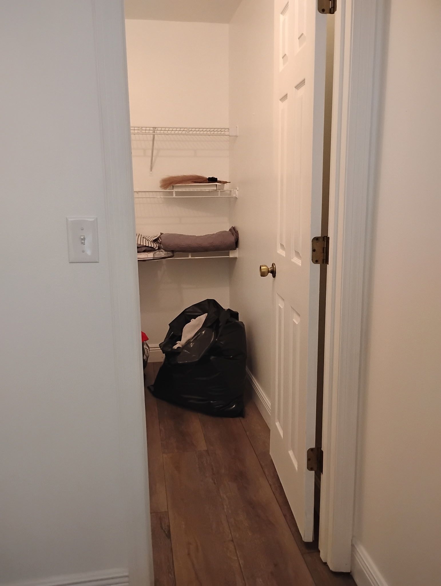 Open closet with shelves, a black bag, and towels inside a white-walled room with wooden flooring.