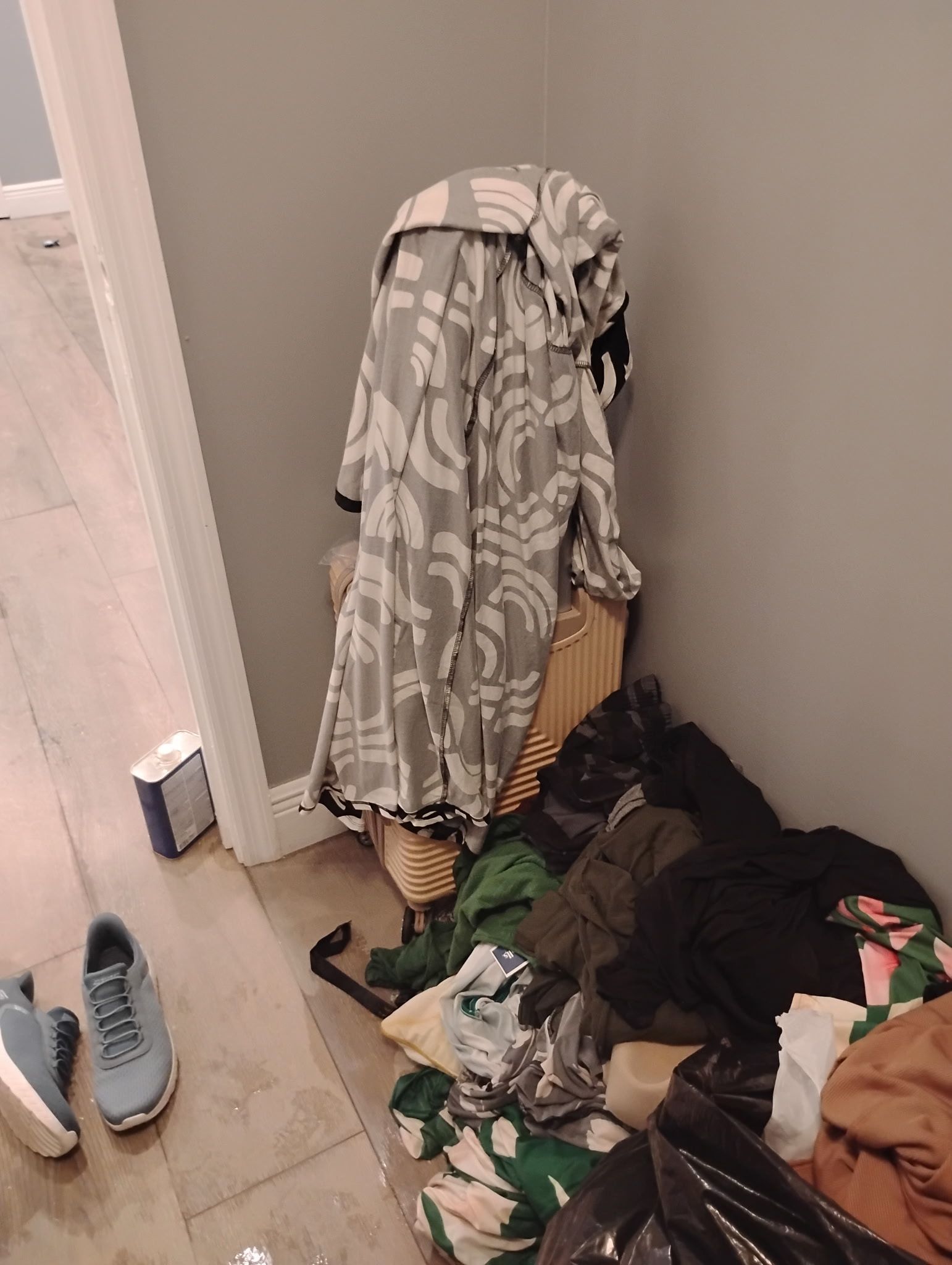 Pile of clothes in a corner of a room, including a floral pattern, with shoes and a small box nearby.