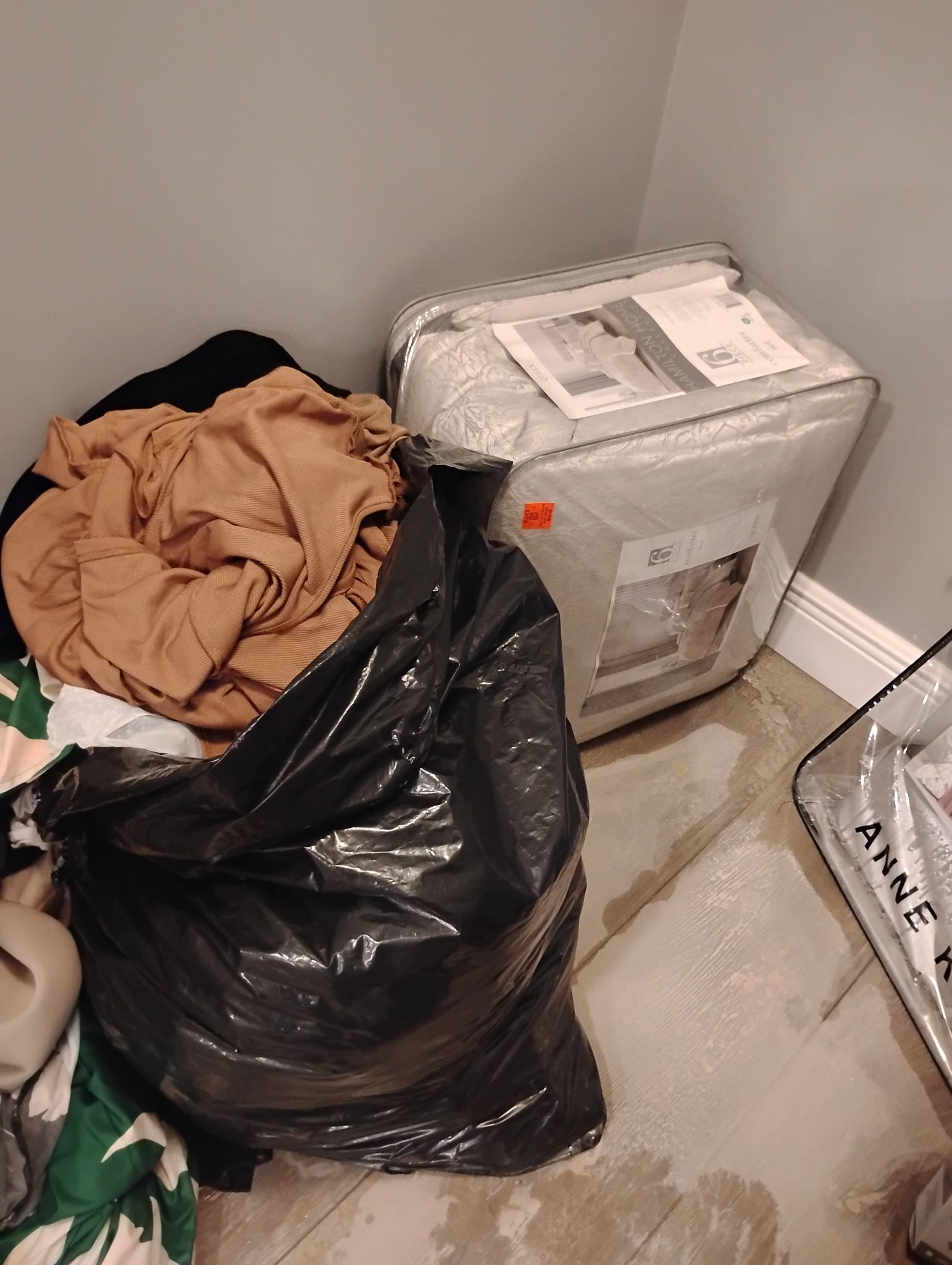 A corner with a black trash bag full of clothes, a packaged comforter, and a white baseboard.