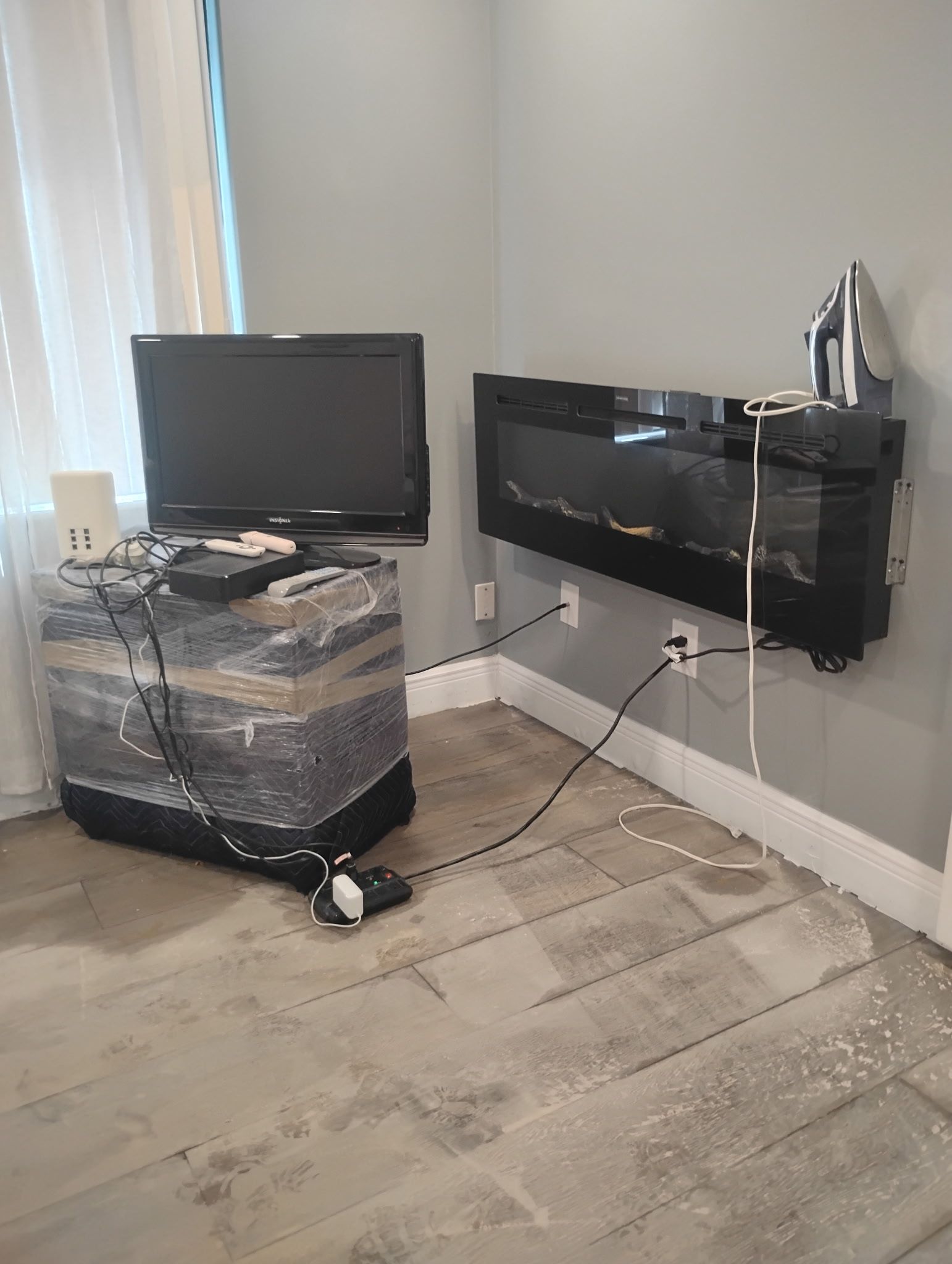 A television atop a wrapped storage container, electric fireplace on the wall, and messy cords in a room with a light floor.