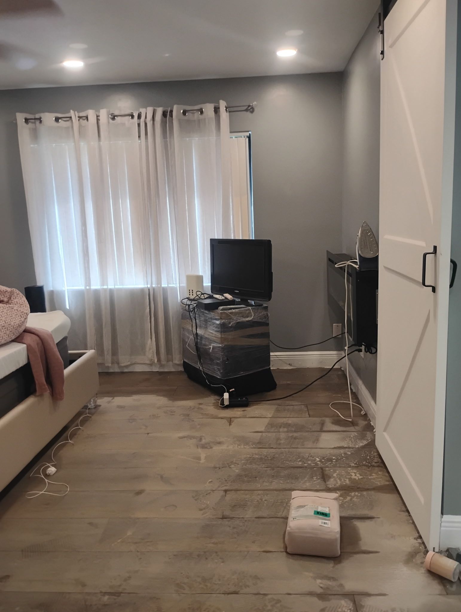 Bedroom with damaged flooring, a window with curtains, and a sliding barn door.