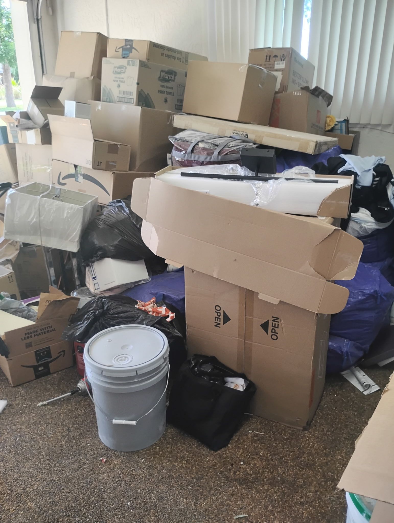 Pile of cardboard boxes, clothing, and other items in a cluttered room. A bucket and a black bag are in the foreground.