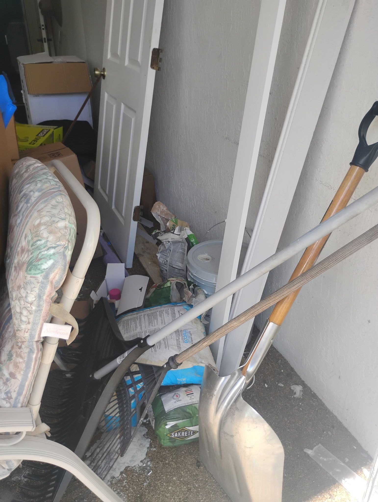 Cluttered storage area with a chair, shovel, and other items against a wall and door.