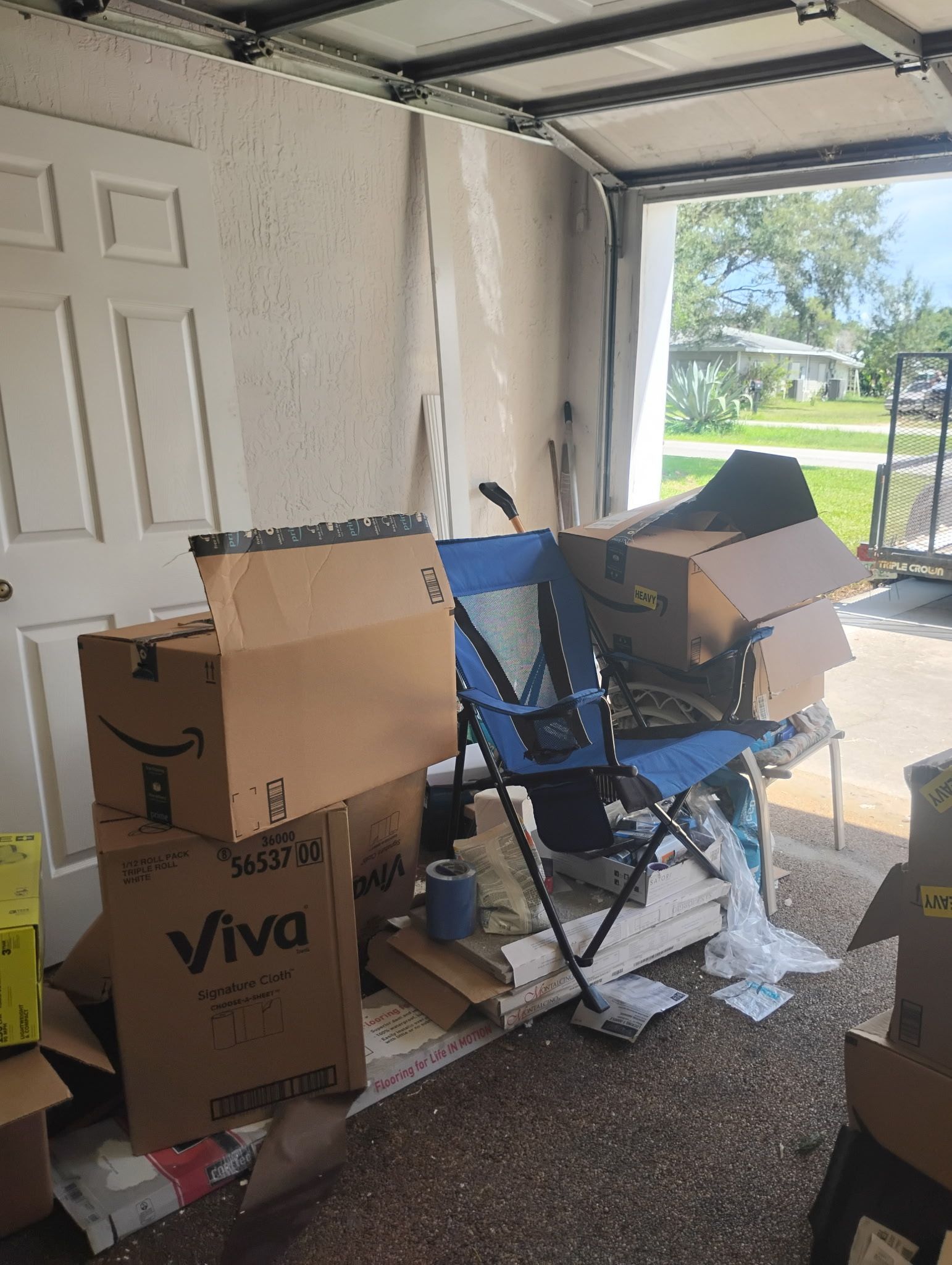Boxes and a folding chair clutter a garage, with the open door revealing a street and green lawn.