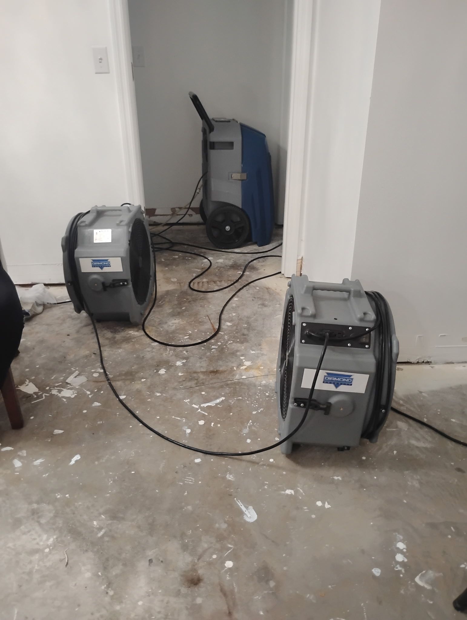 Three industrial fans and a dehumidifier drying a room with white spattered concrete floor and doorway.