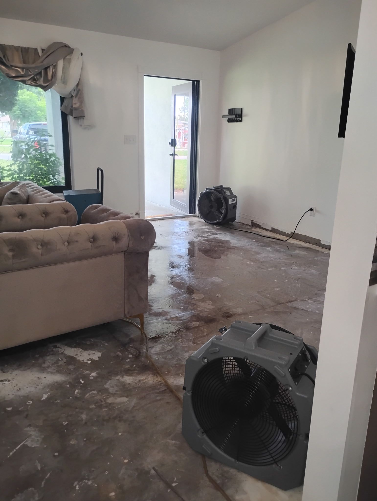 Flooded room with fans. Beige sofa on the left, open door, water on floor, white walls.