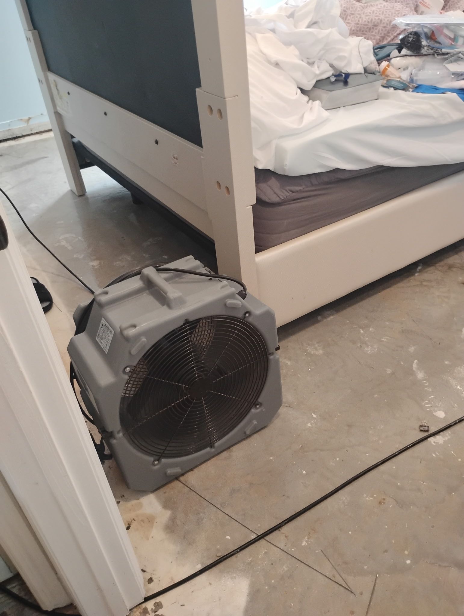 A gray industrial fan on a concrete floor next to a white bed frame with bedding.