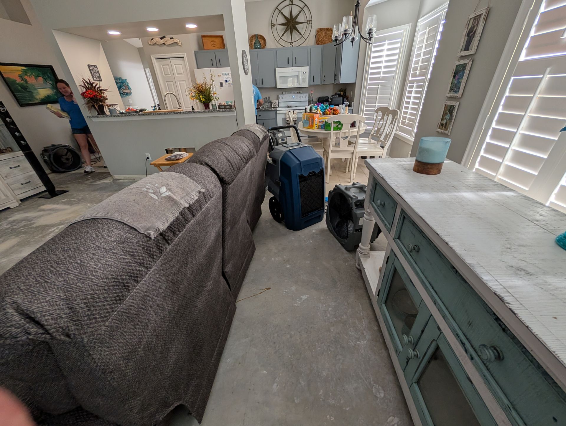 Living room with couch, rug cleaner, and kitchen visible. Open shutters let in light.