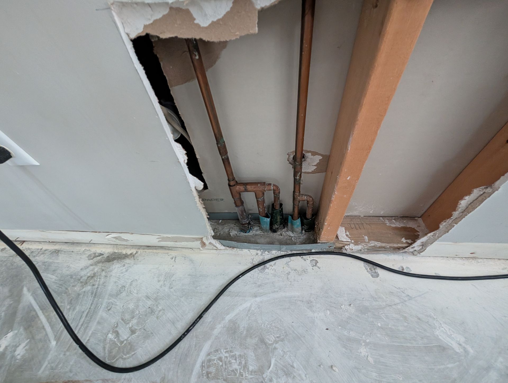 Hole in ceiling reveals copper pipes and plumbing. Gray drywall, wooden beams, black cable visible.