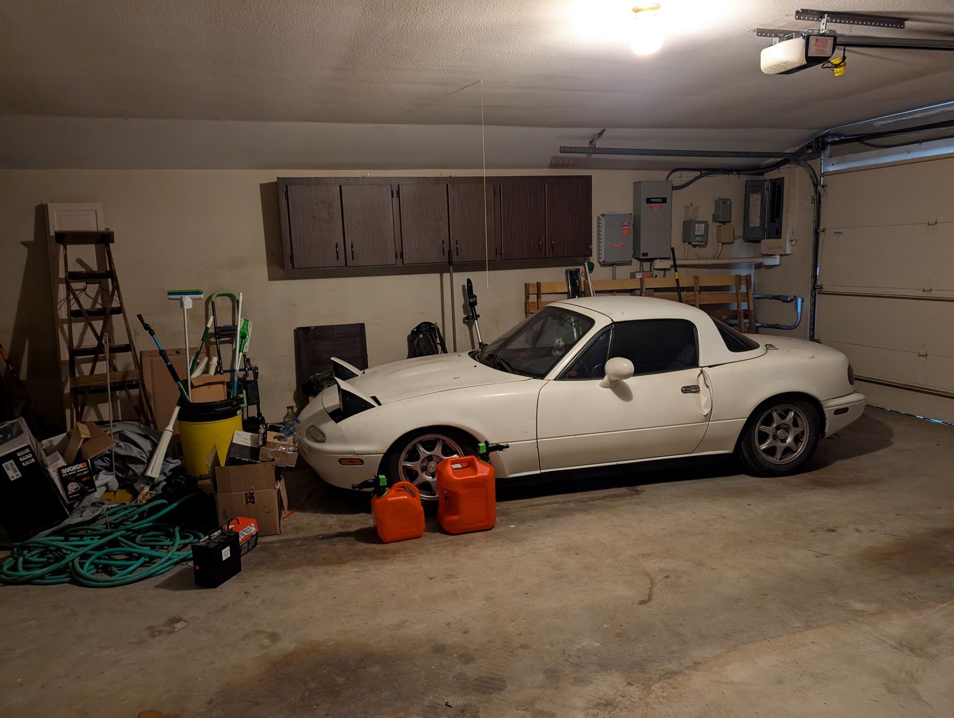 White Mazda Miata in a cluttered garage, with two gas cans in front.