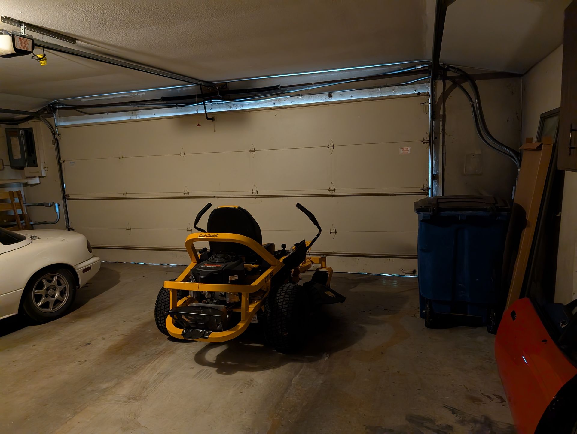 Garage interior: yellow riding mower, white car, blue trash bin.