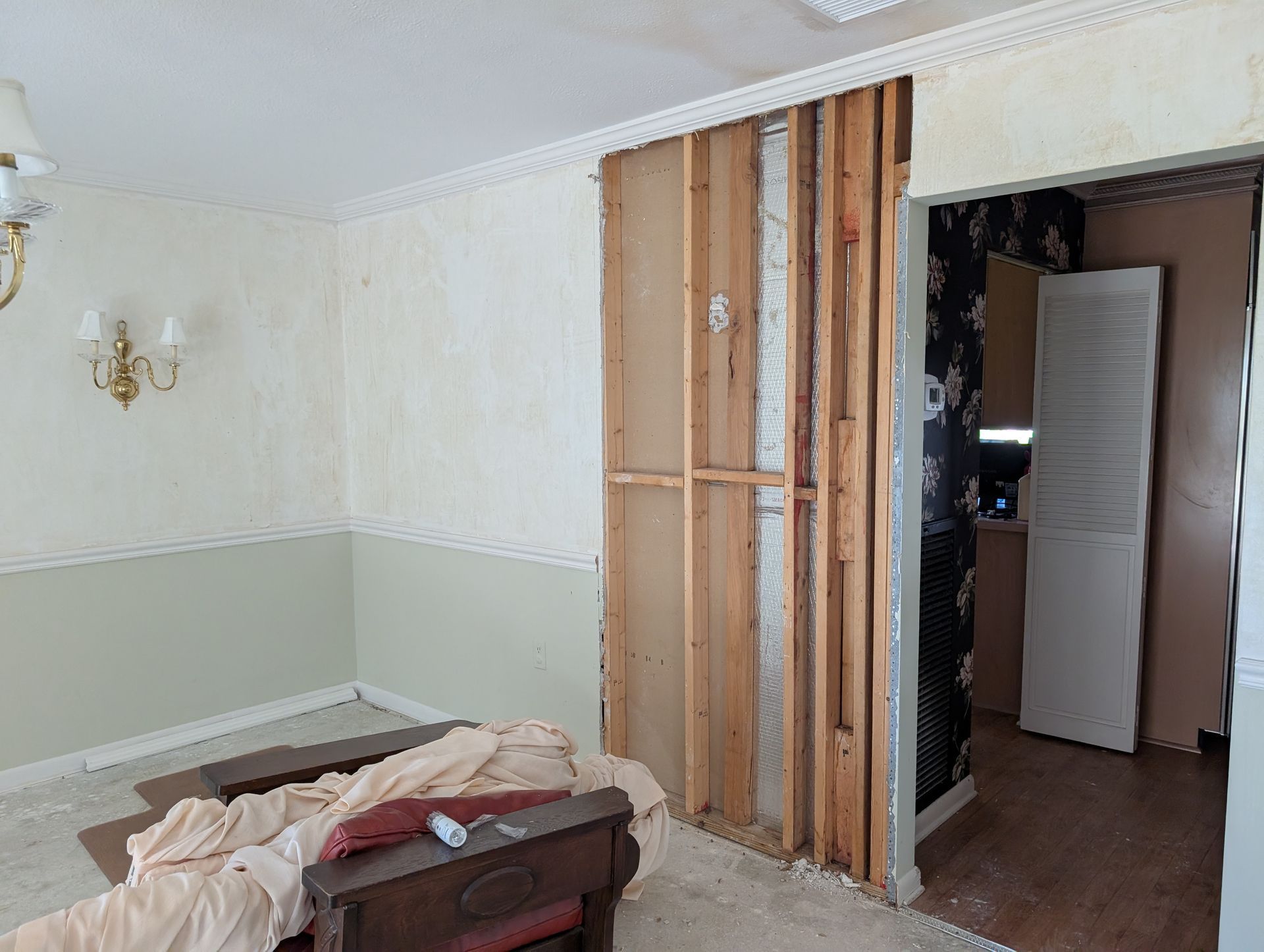 Interior room undergoing renovation; exposed wall studs, floral wallpaper, and partially painted walls.