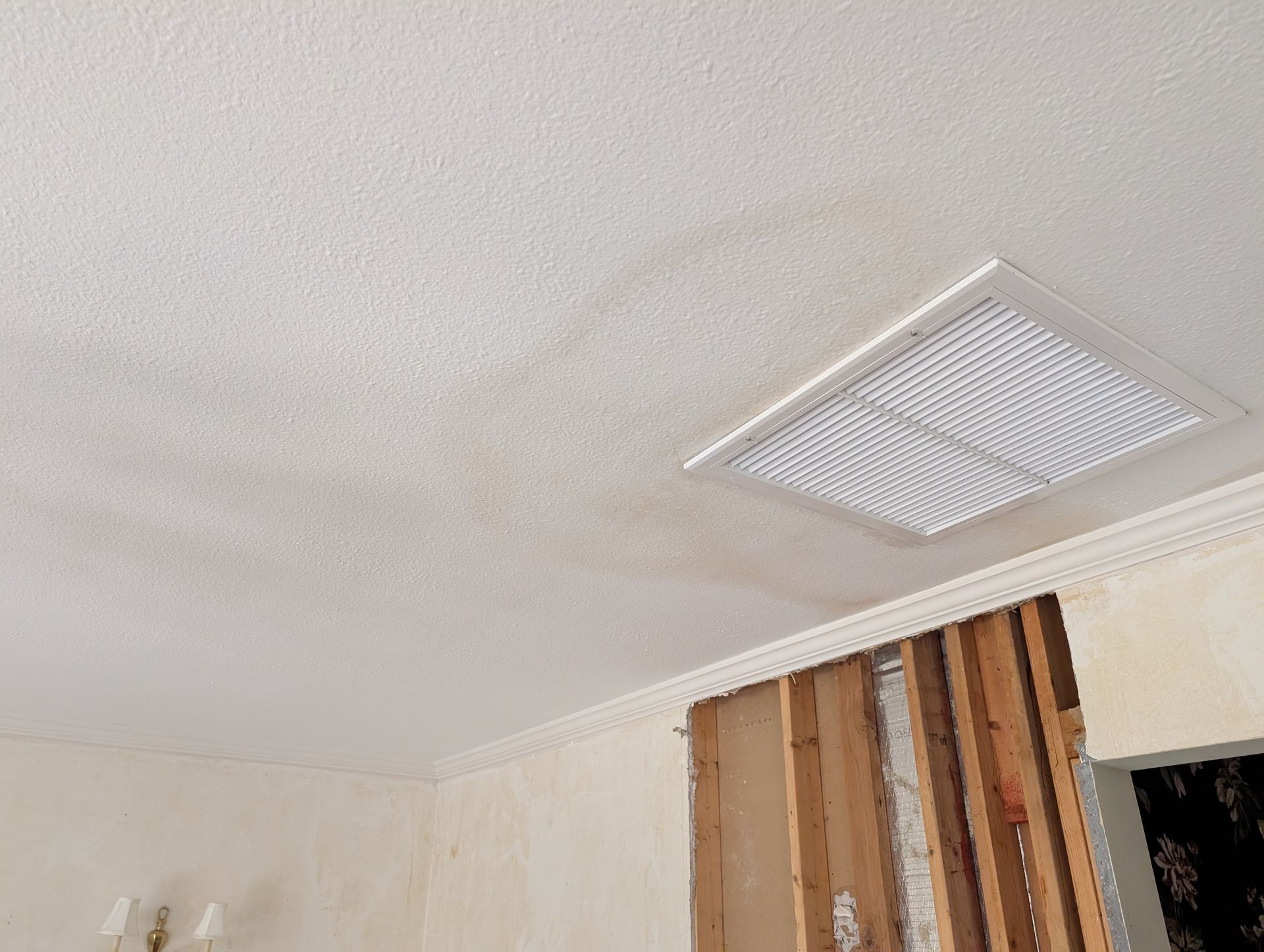 Ceiling with water damage and a square air vent; exposed wall studs.