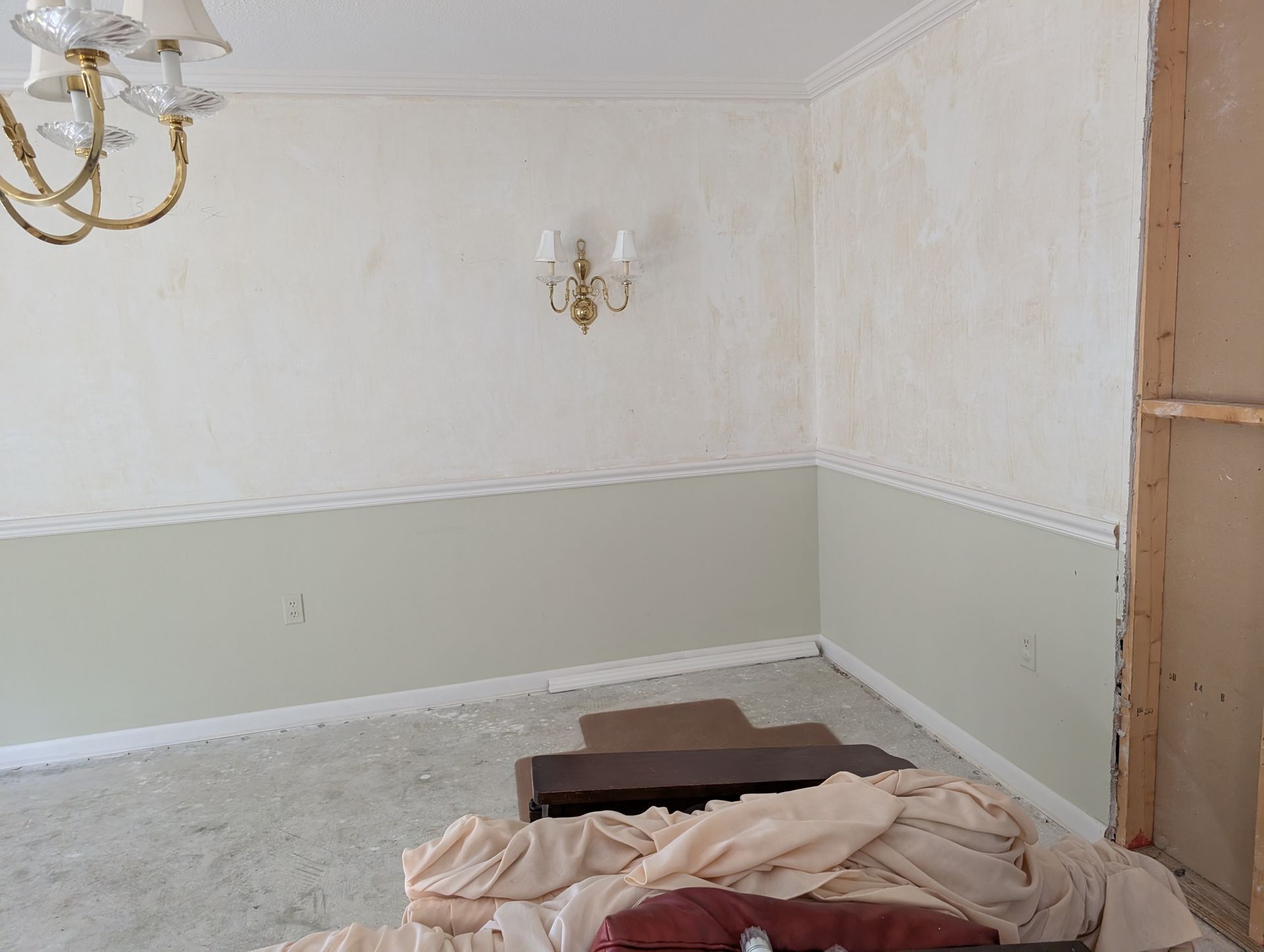 Room interior under renovation: walls with two-tone paint, a chandelier, and debris on the floor.