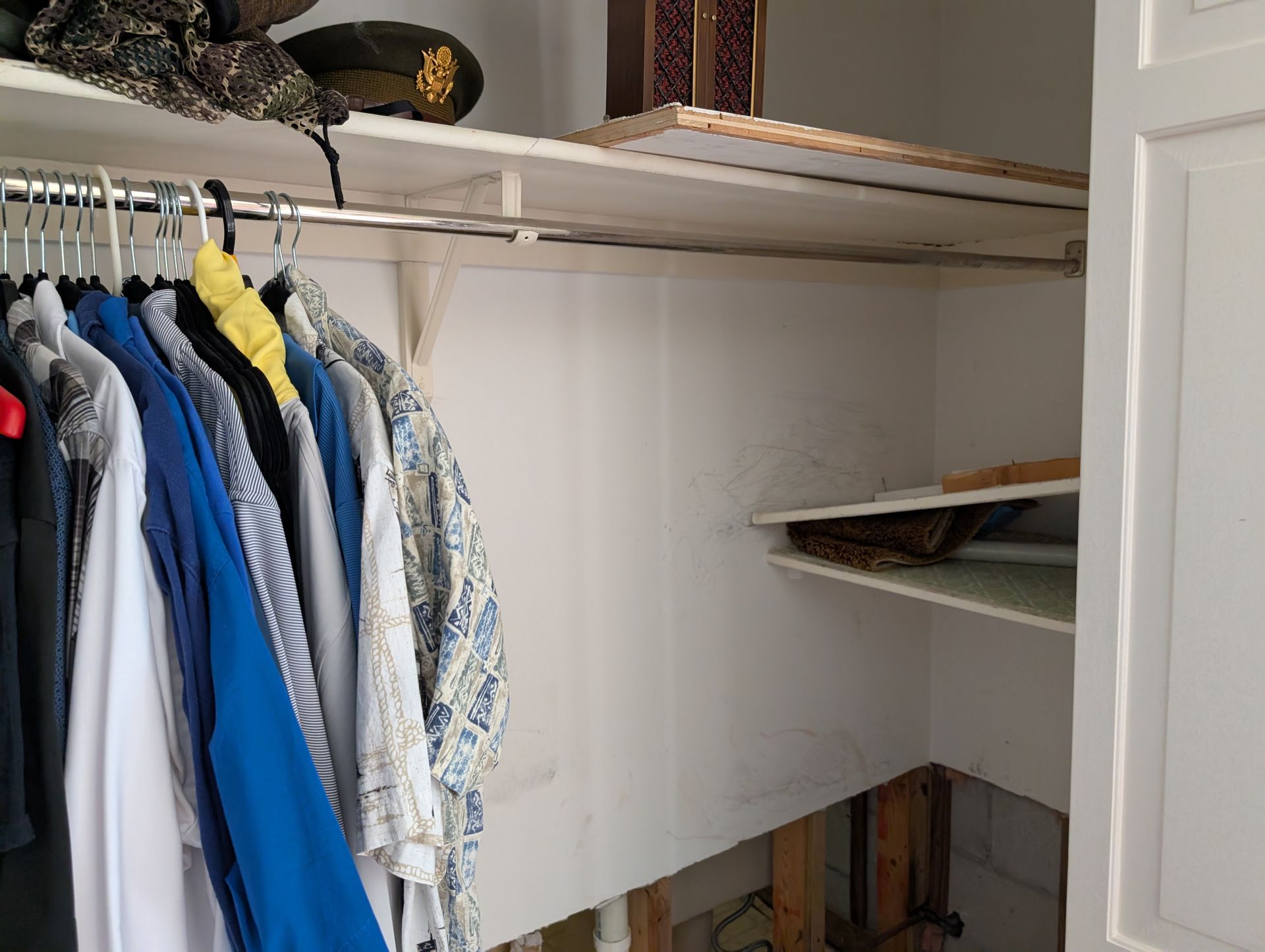 Closet with clothes hanging on a rod and shelves with items stored above a space where the flooring is missing.