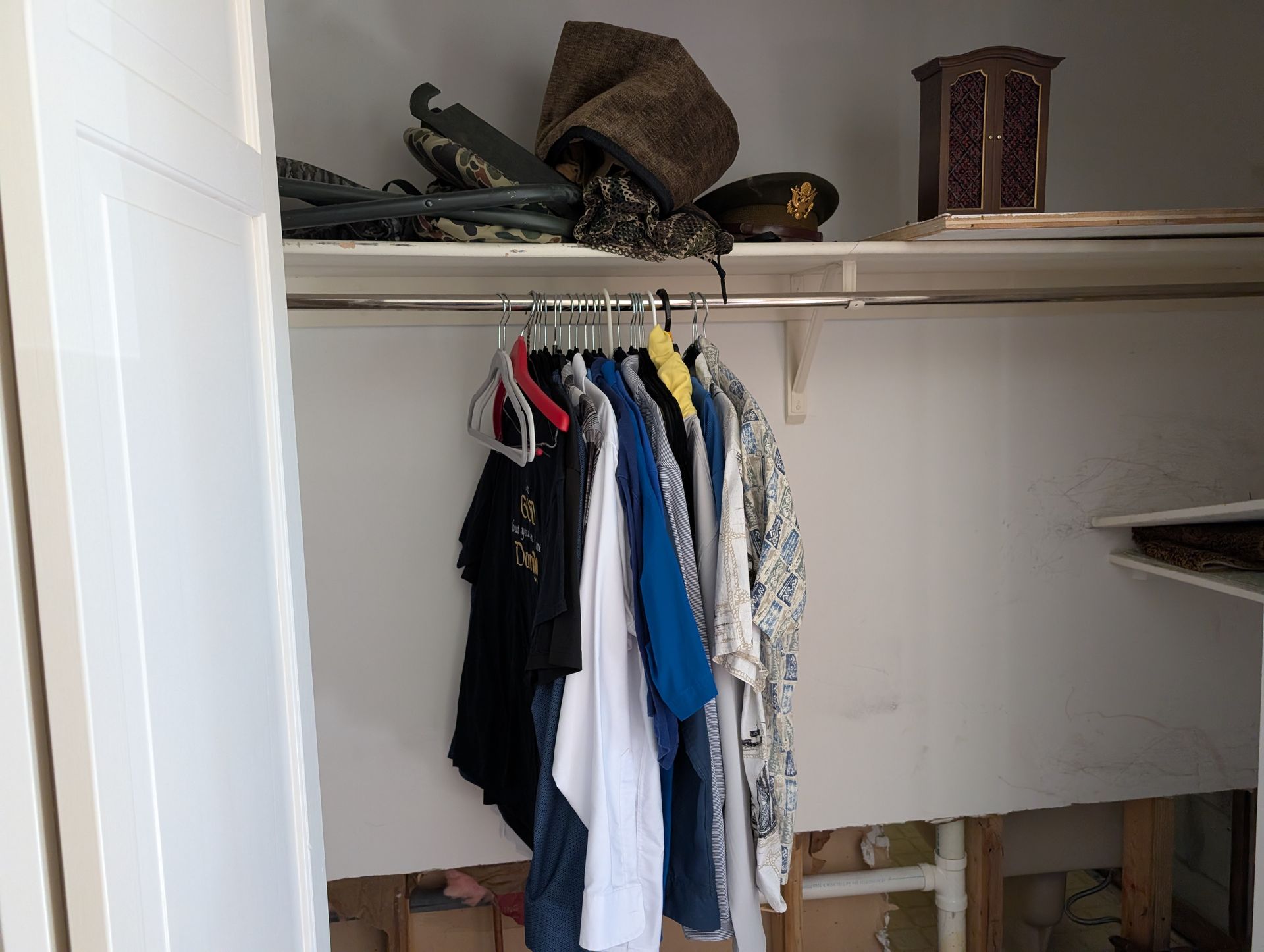 Inside a closet: clothes hanging on a rod, shelf with hats and a small decorative box. The closet wall is damaged.