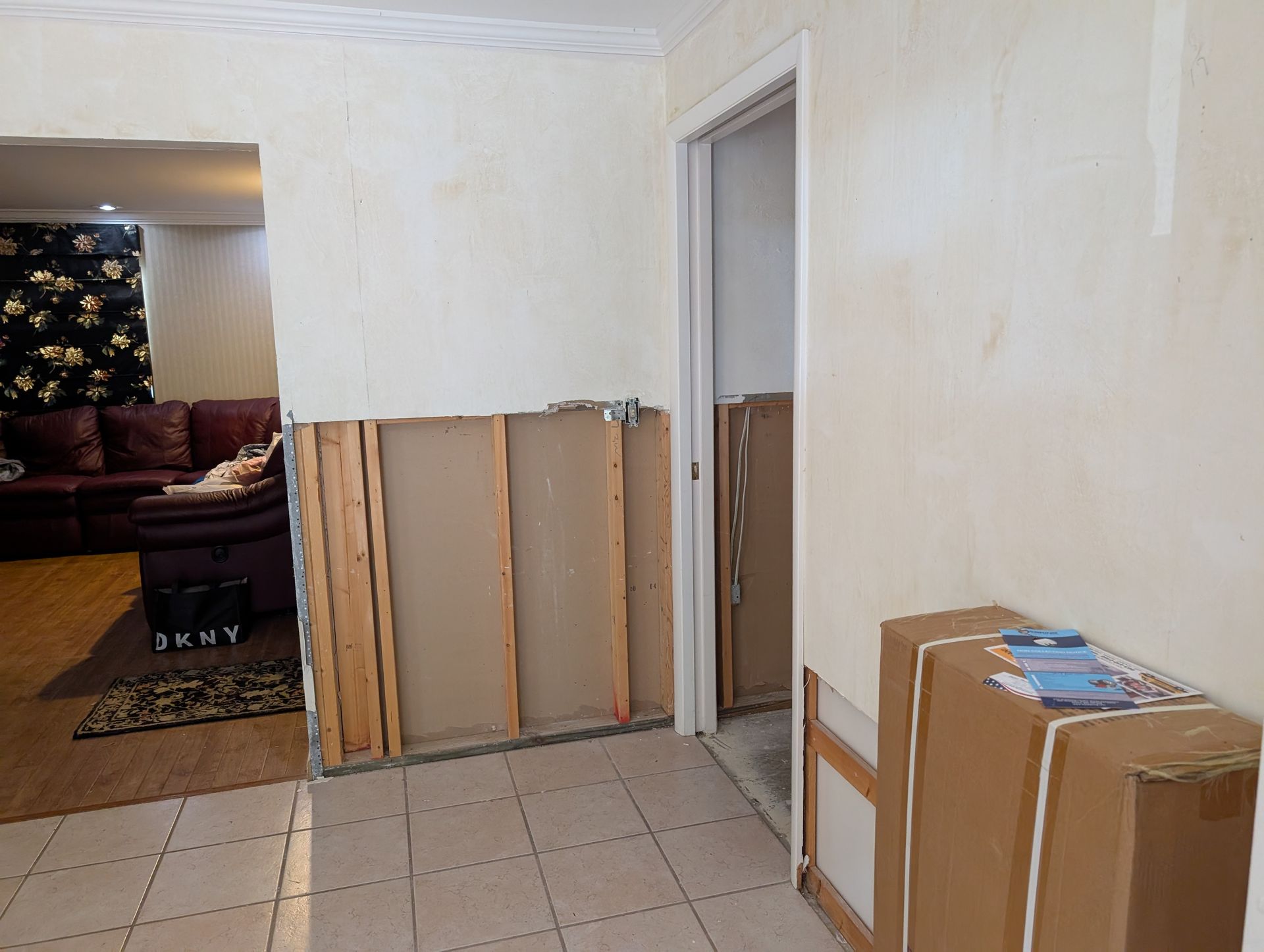 Interior view of a hallway with damaged drywall, open doorway to living room, and cardboard boxes.