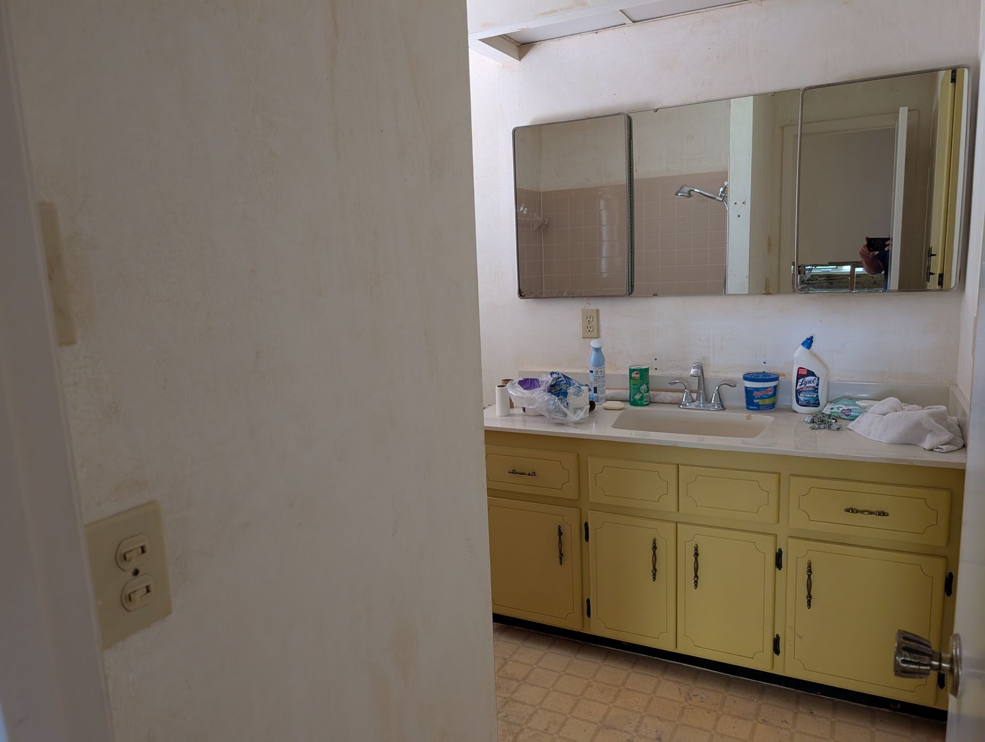 Bathroom with yellow cabinets, three-door mirrored medicine cabinet, sink, and various cleaning supplies.
