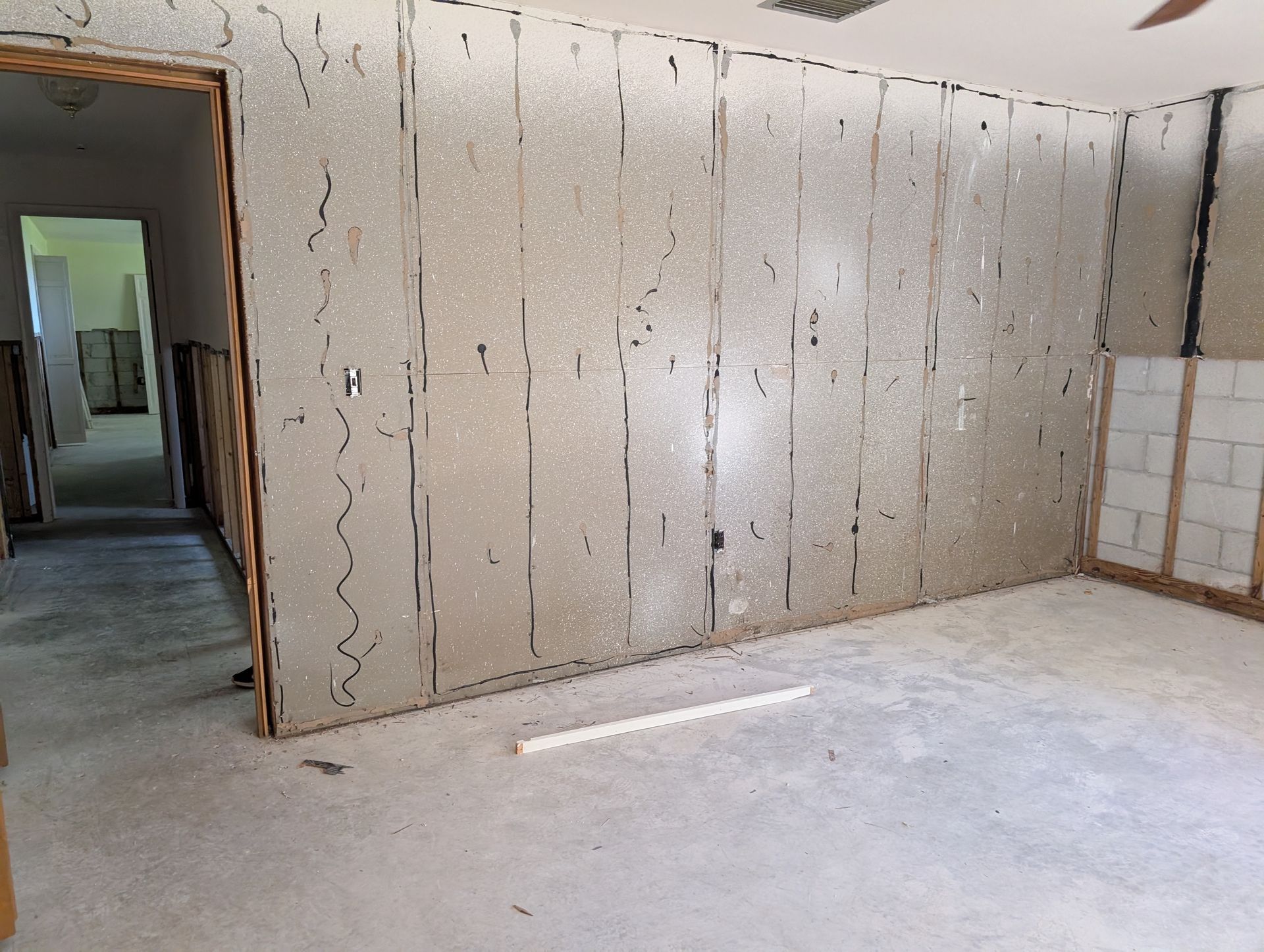 Interior room under construction, with exposed wall panels and a concrete floor. A doorway leads to another room.