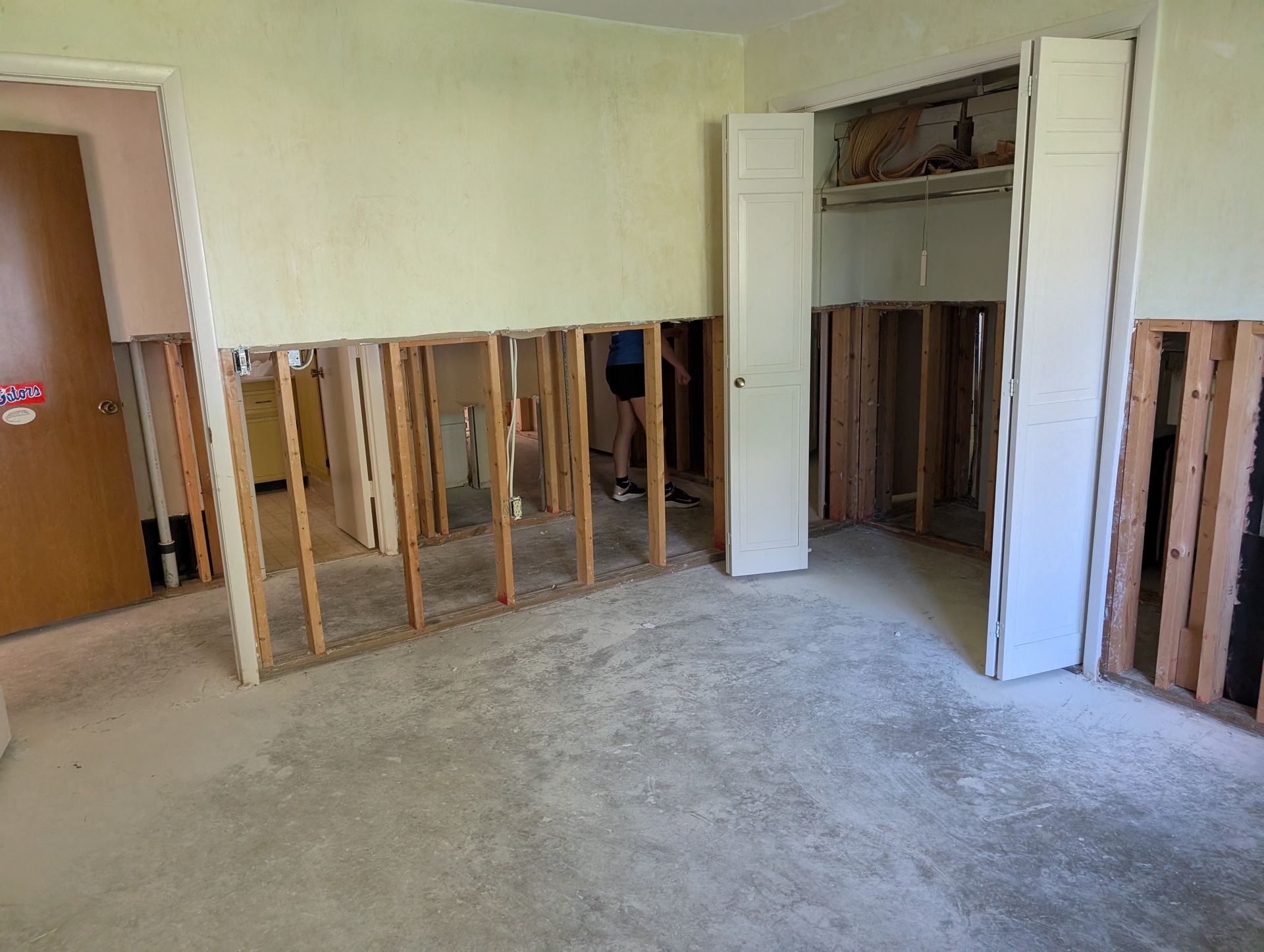 Room gutted, walls open to studs, with cabinets standing. Person in doorway.