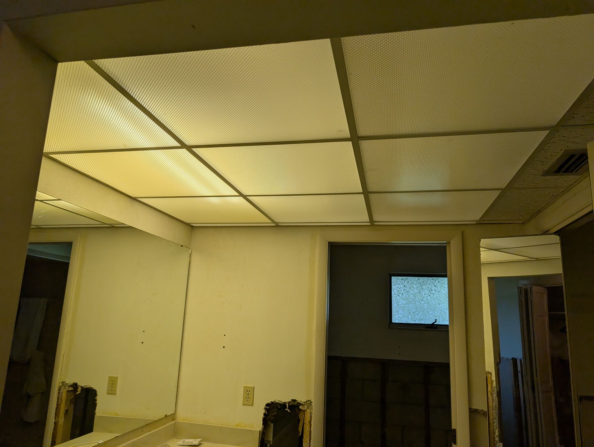 A bathroom interior with a drop ceiling, mirror, and open doorways, illuminated by overhead lighting.