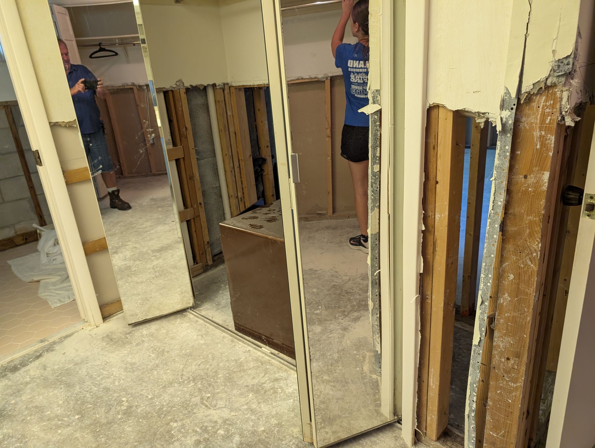 People removing mirror from a wall; interior view of room with exposed studs, debris on floor.