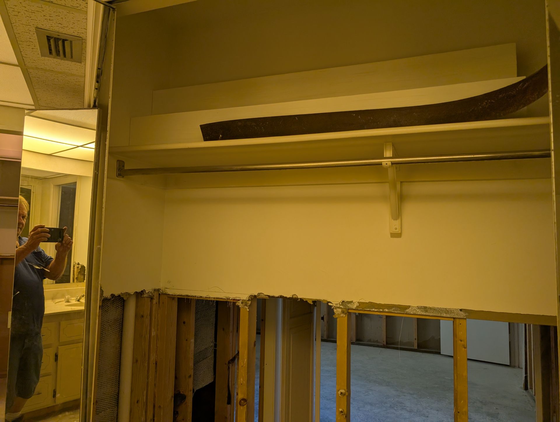 A person takes a photo of an open closet with drywall removed, revealing the wooden studs and a partially visible bathroom.