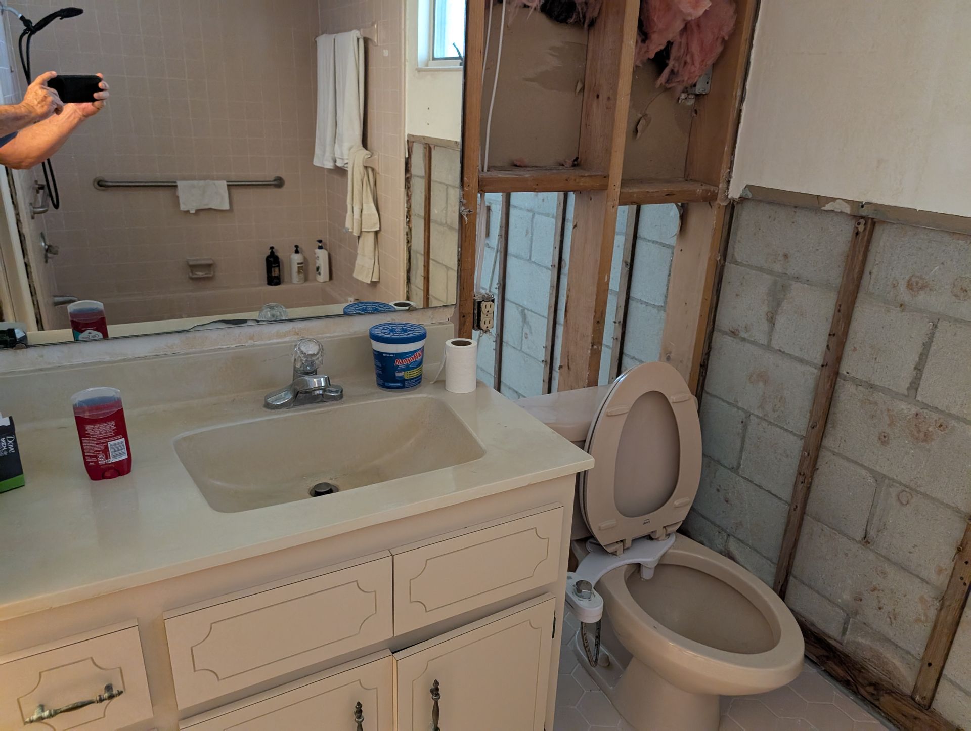Bathroom renovation in progress, damaged wall, exposed studs, toilet, sink, mirror.