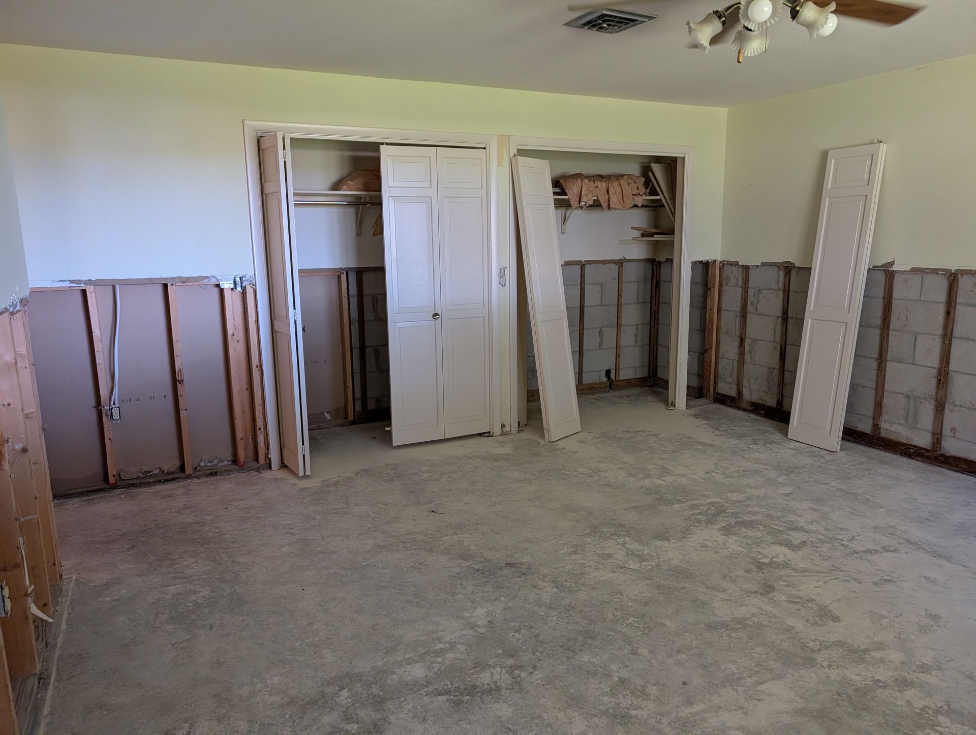 Room under renovation; drywall and closet doors removed, concrete floor exposed, light-colored walls.