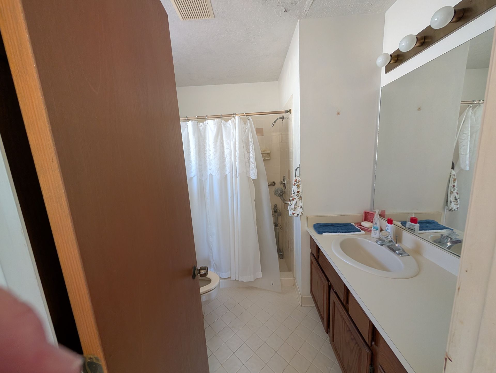 Bathroom with shower, vanity, and open door. White walls, tile floor, brown cabinets.