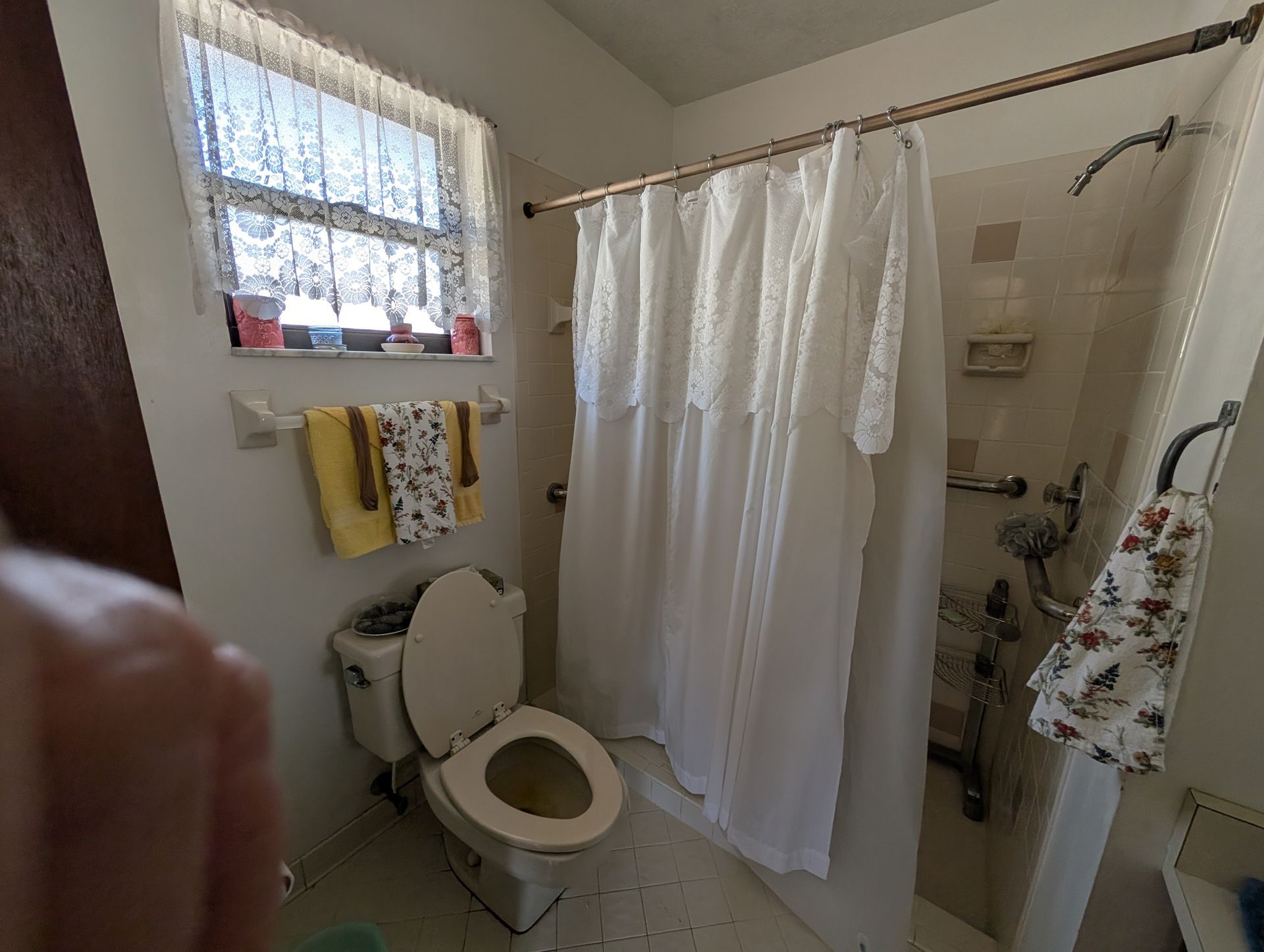 Bathroom with toilet, shower, and window with curtains; towels hang on a rack.