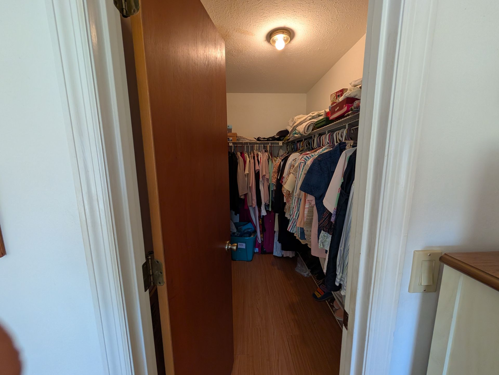 Closet with clothes on racks and ajar brown door. White trim, wood flooring.