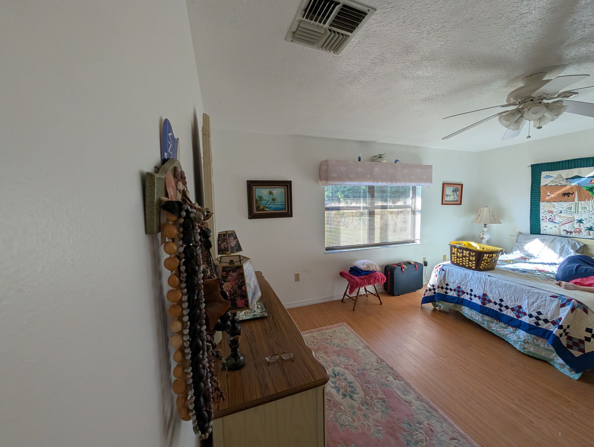 Bedroom with wood dresser, bed, window, and ceiling fan. Light-colored walls and wood-look flooring.
