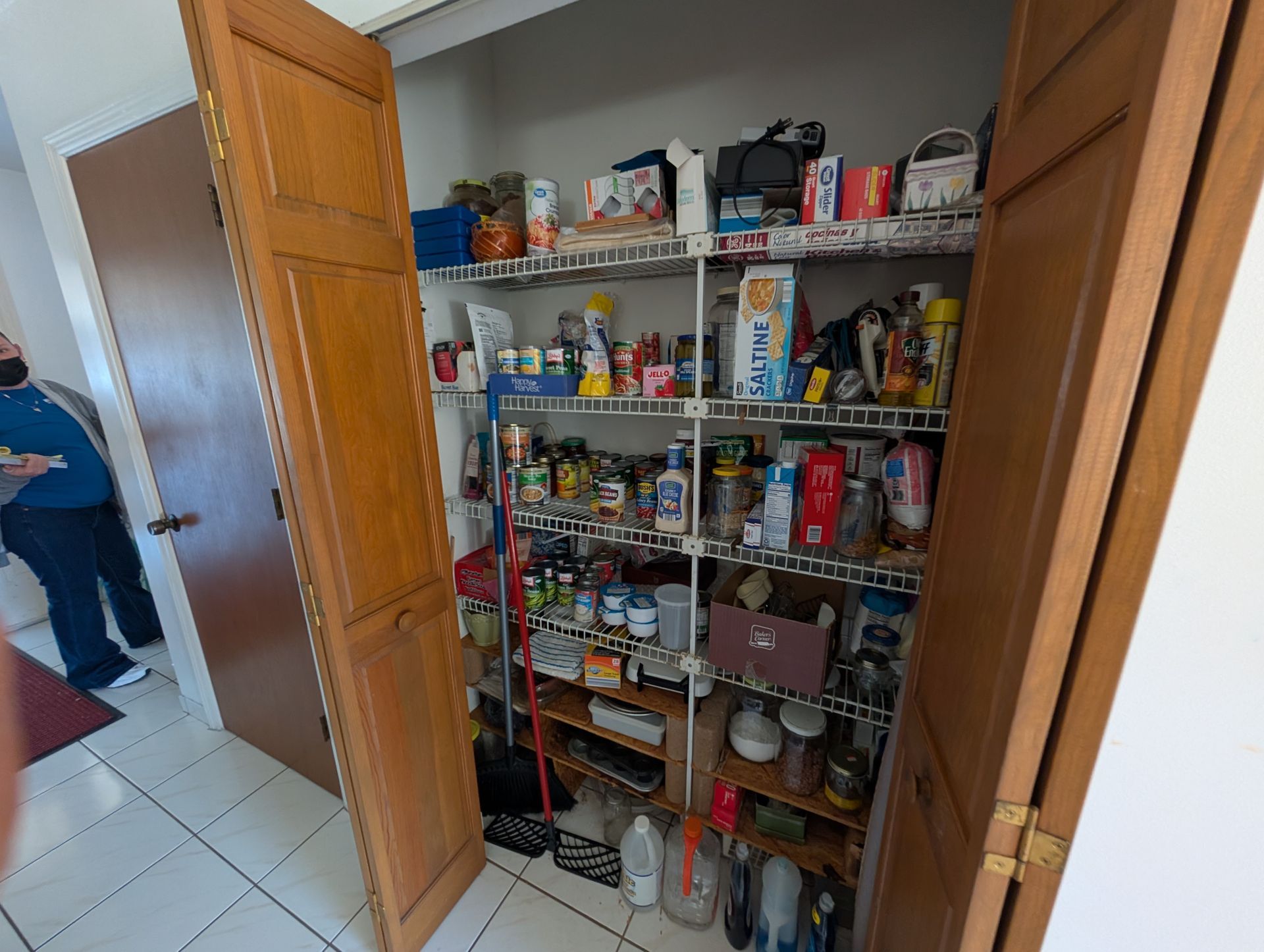 A pantry with wooden doors open, filled with various food items and cleaning supplies on wire shelves.