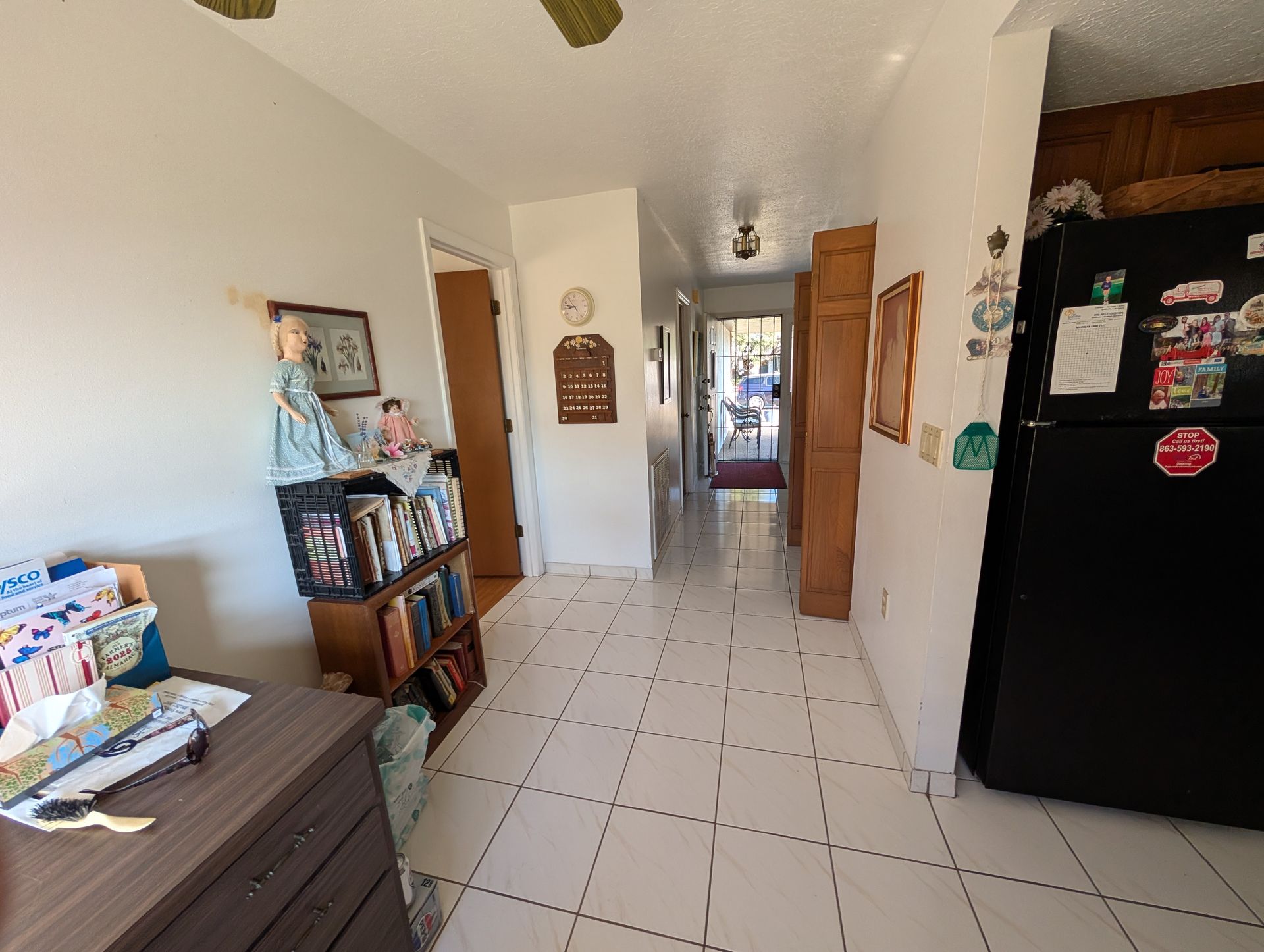 Hallway with white tiled floor, dark refrigerator, bookshelf, and doorway to the outdoors.