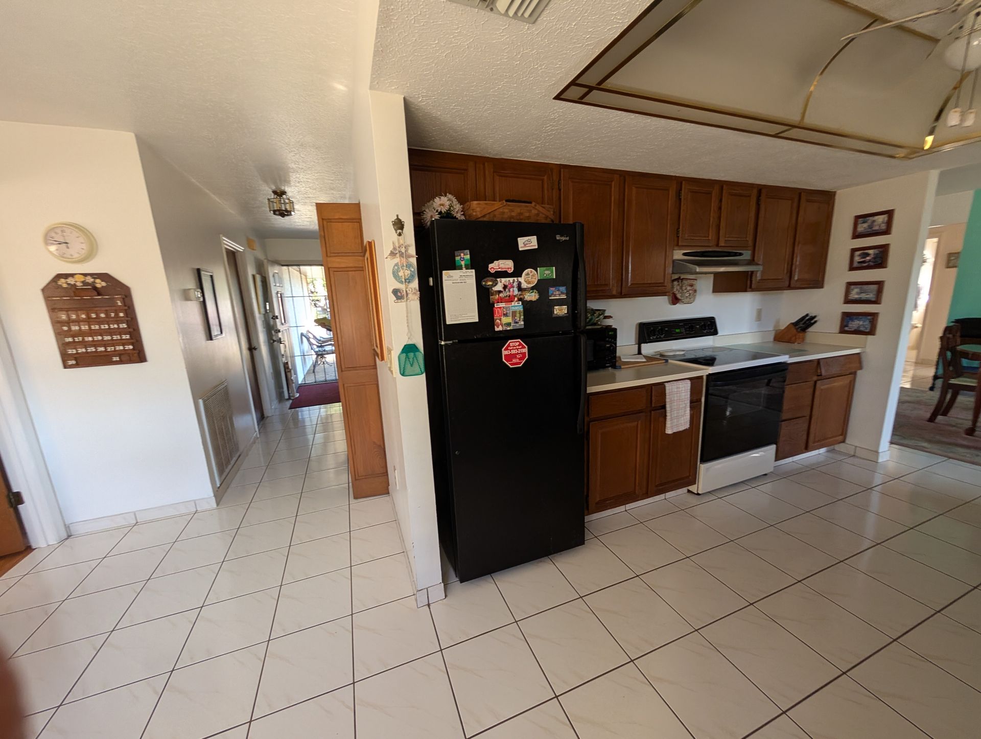 Kitchen with brown cabinets, black refrigerator, and white tiled floor. Hallway visible.