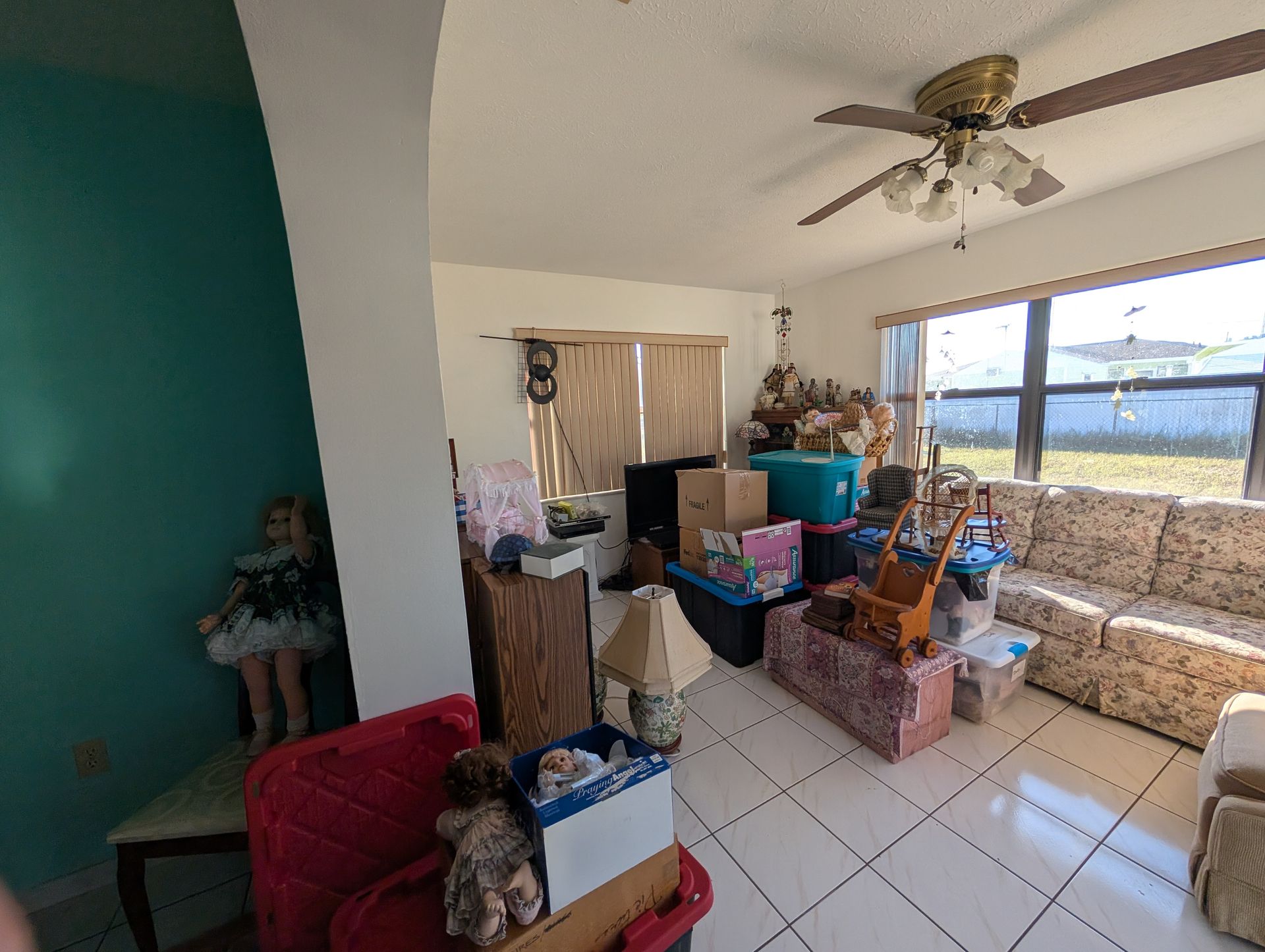 Cluttered living room with boxes, furniture, and a doll. White walls, tile floor, and a ceiling fan.