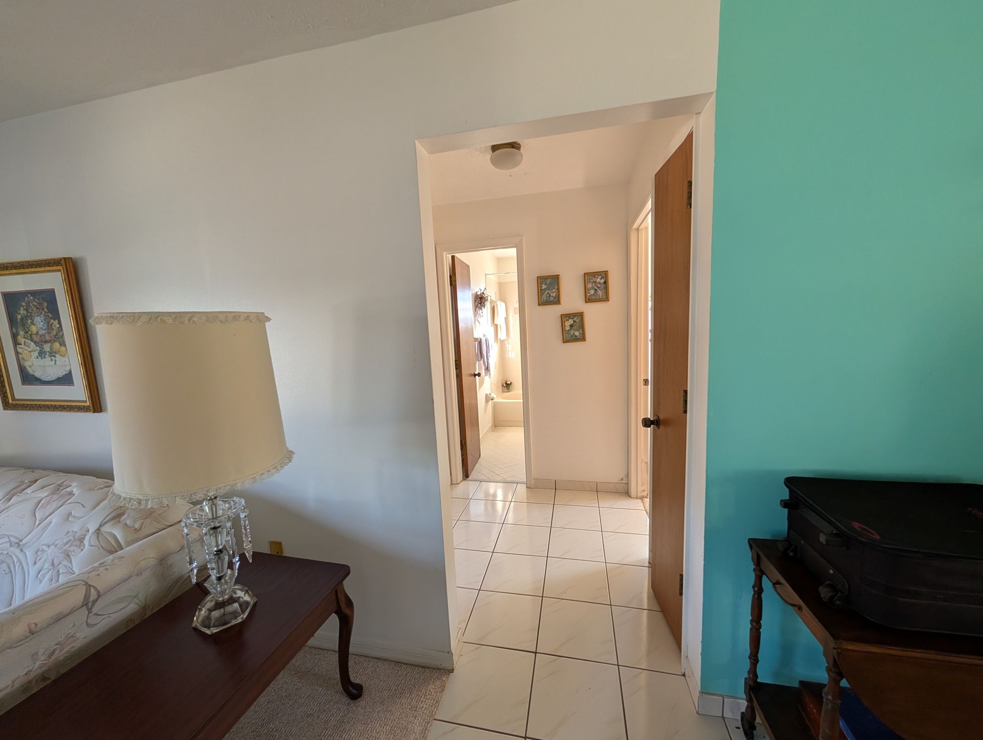 Bedroom doorway leading to a hallway with a bathroom visible. White walls, turquoise accent wall, and tiled floors.