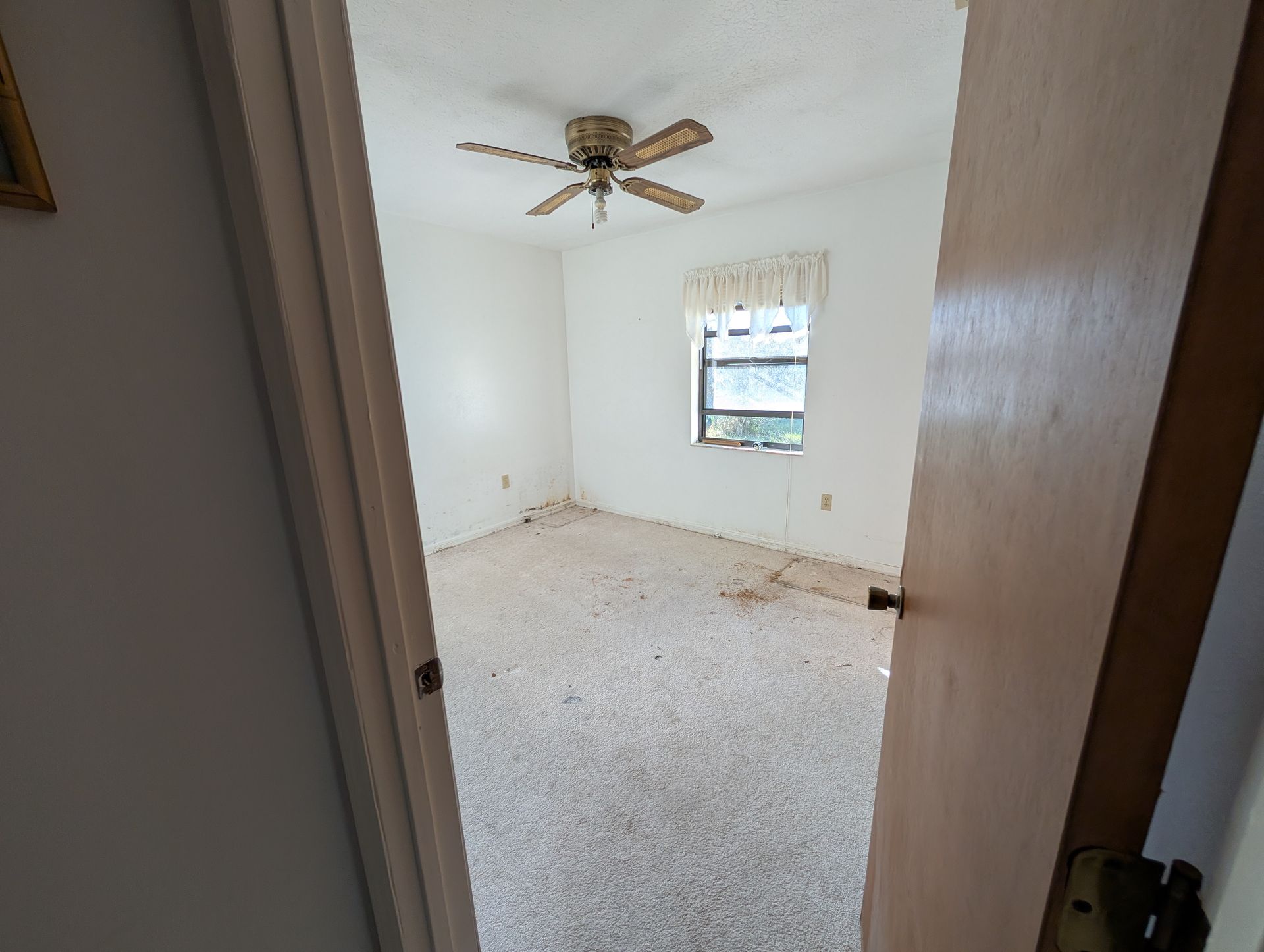 Empty bedroom with white walls, ceiling fan, and speckled flooring seen through a doorway.