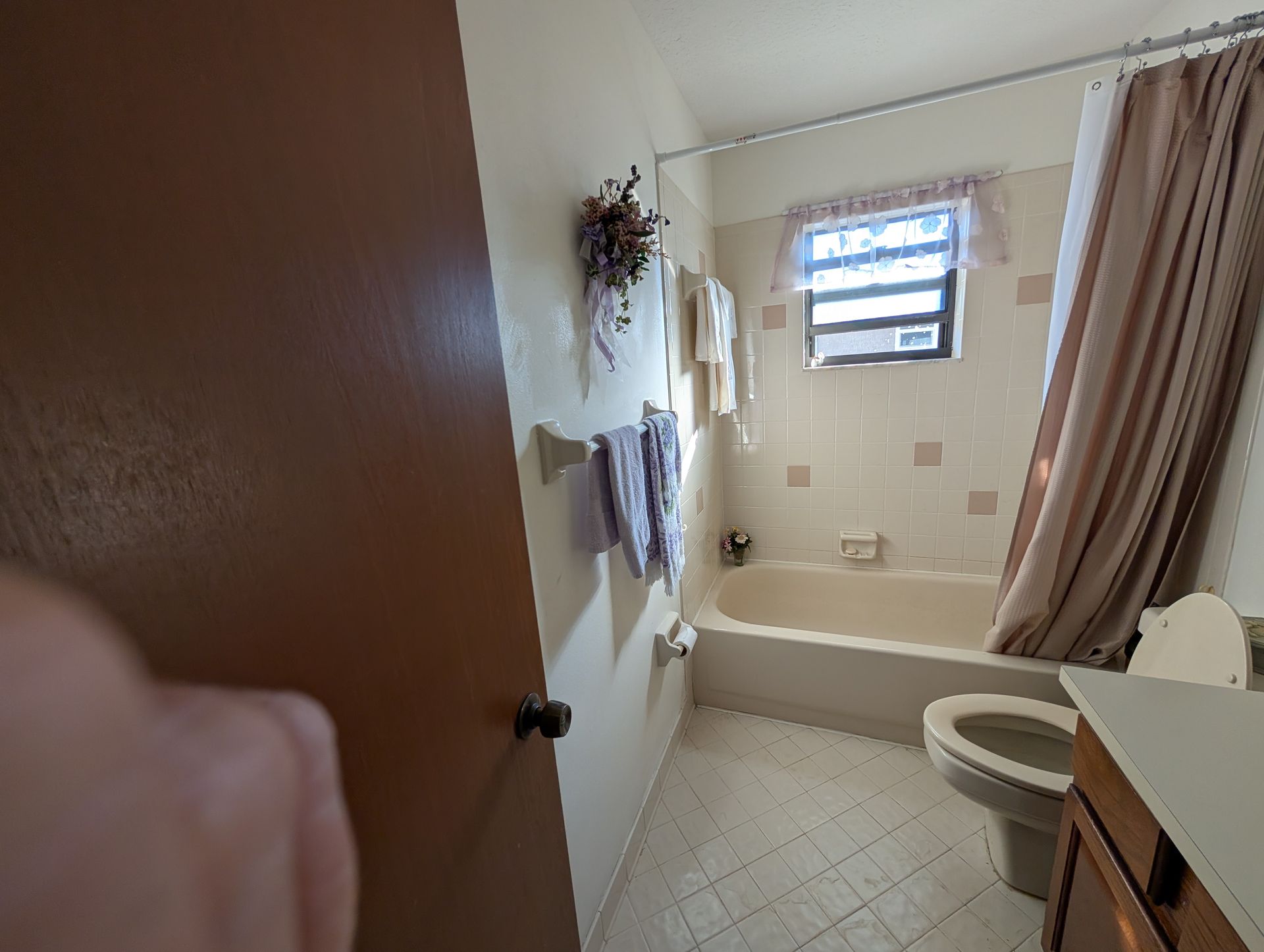 Bathroom with a white bathtub, toilet, and beige shower curtain. A small window with a curtain is above the tub.