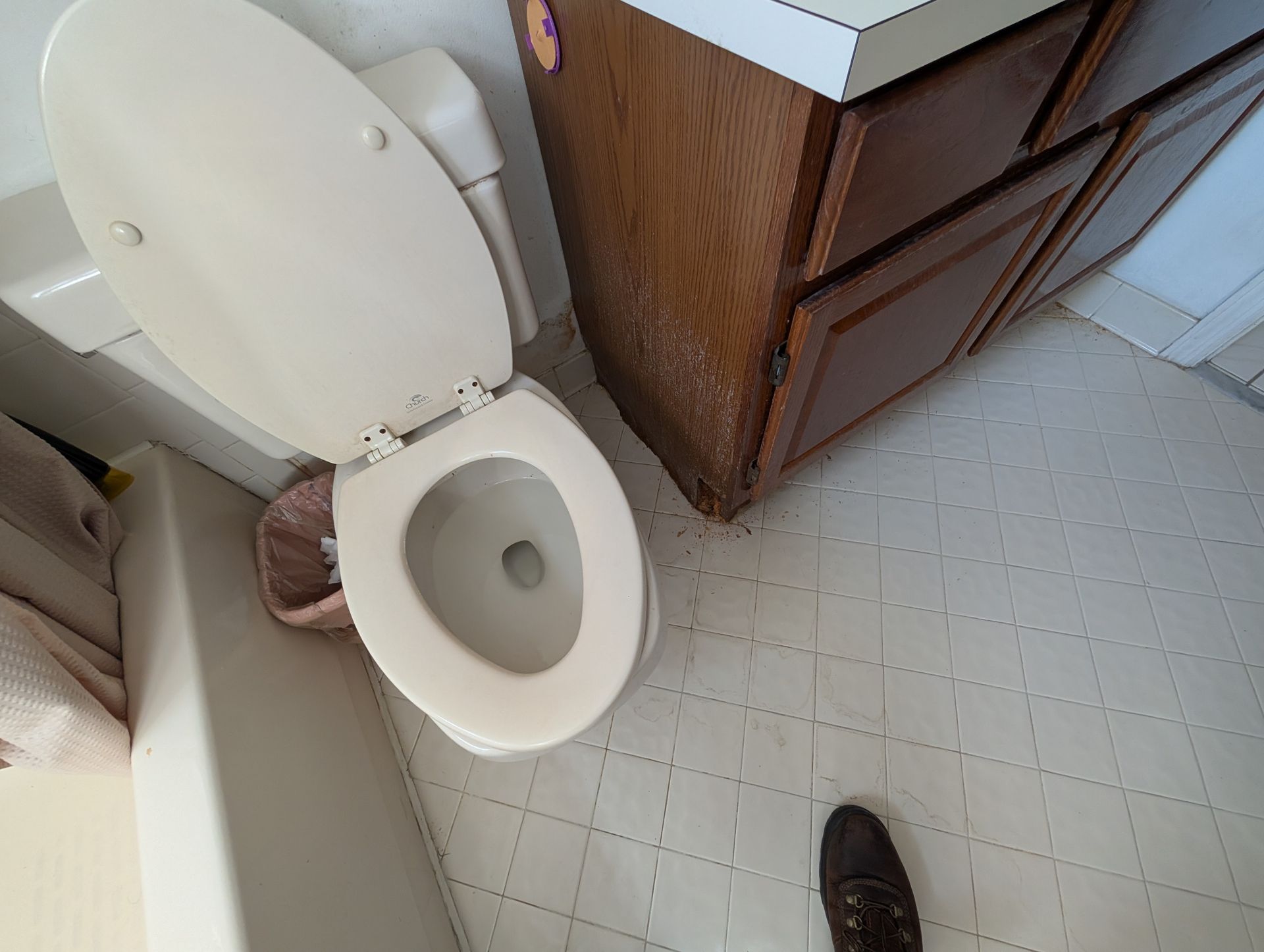 Toilet with open lid beside wooden cabinet, on white tiled floor. A foot in a brown shoe is visible.