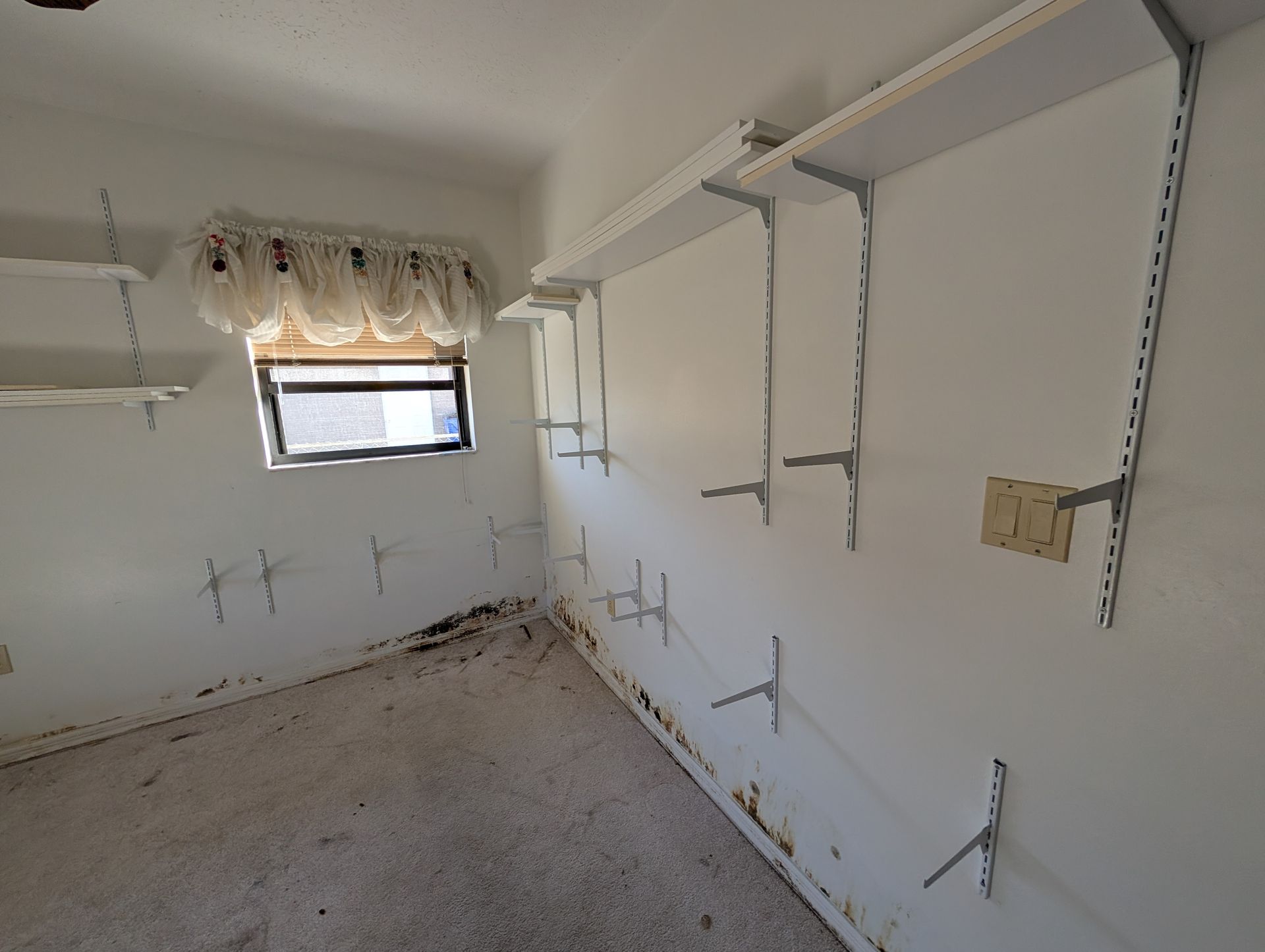 Empty room with white walls, shelves, and window; debris on the floor and visible wall damage.