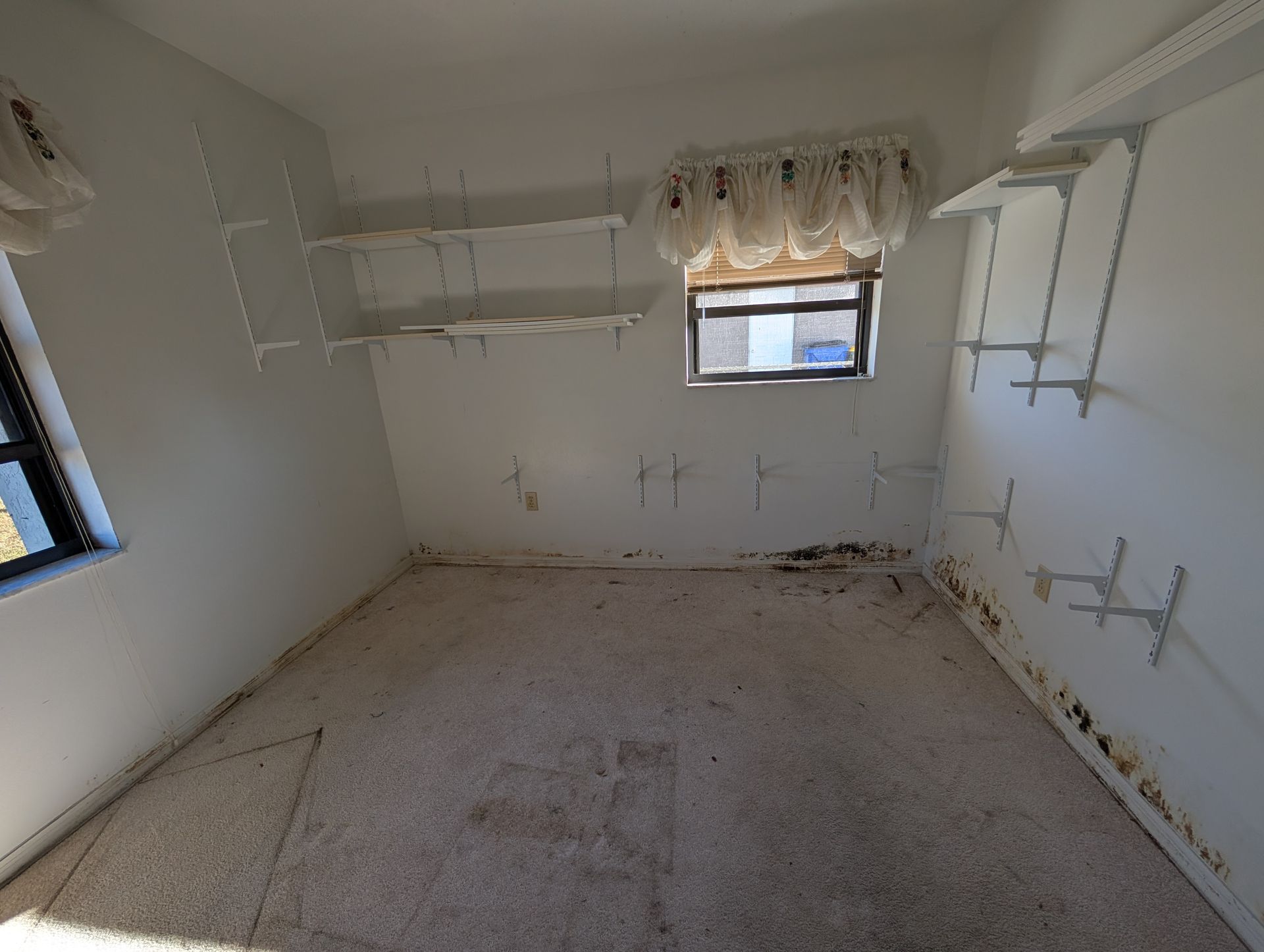 Empty white room with shelving brackets, small window, and stained walls near the floor.